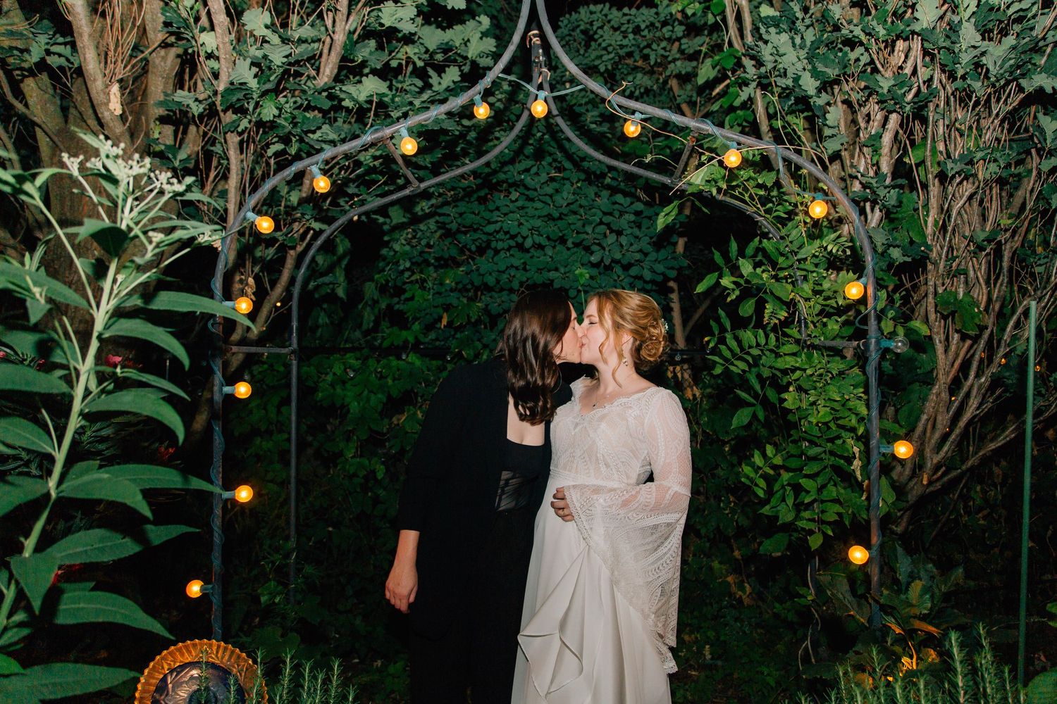 Lesbian couple sharing a kiss under a floral arch with string lights at night.