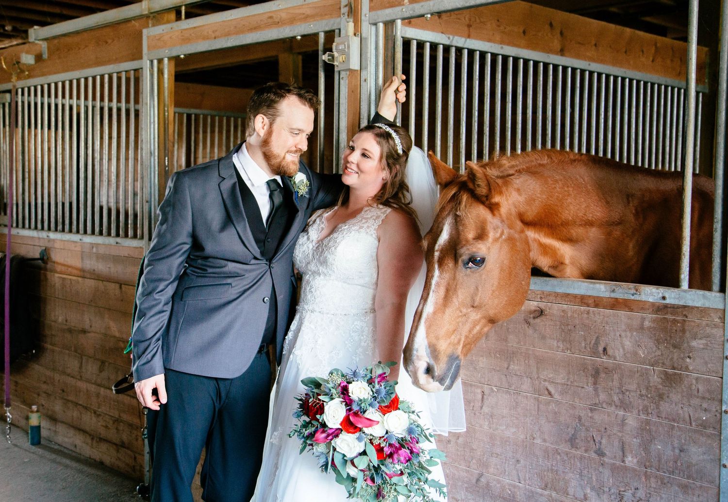 Bride and groom pose with chestnut horse inside rustic barn stables on their wedding day.