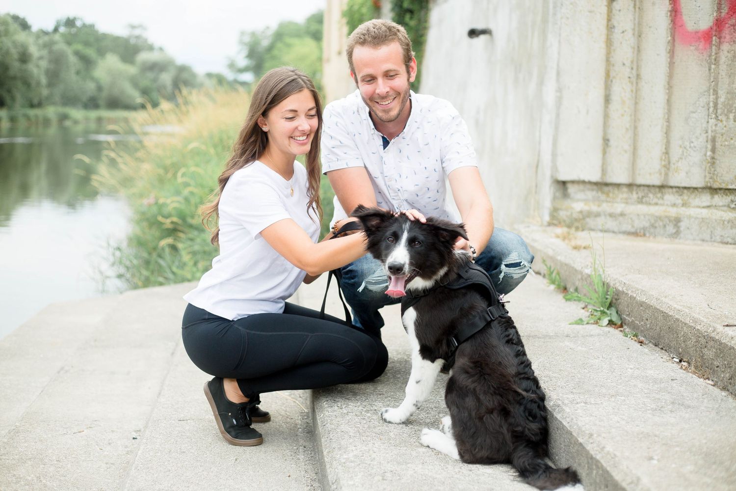 A smiling couple crouches down to pet their black and white Border Collie dog by a riverside.