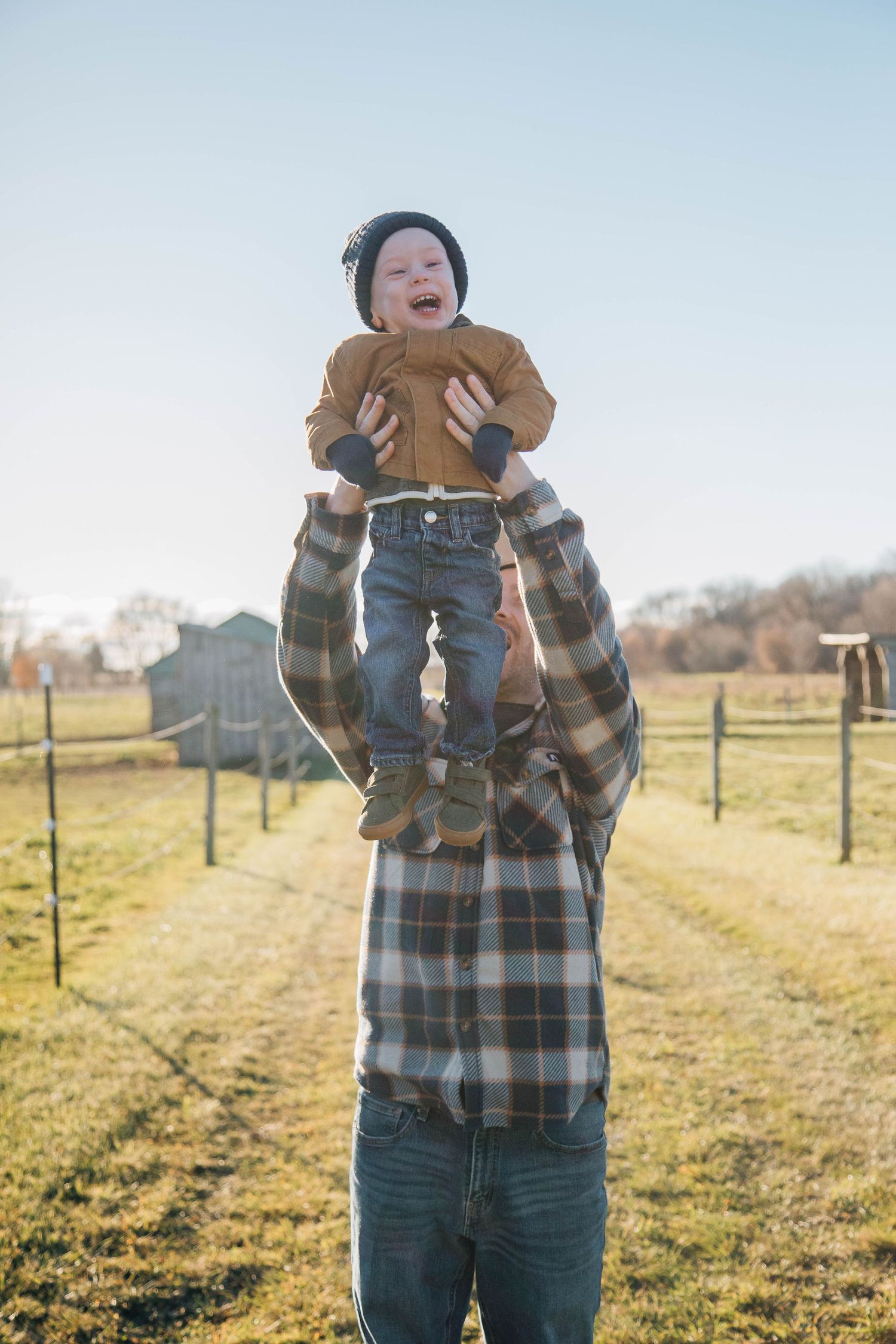 A father joyfully lifts his laughing baby high in the air during an outdoor family photo session on a farm.