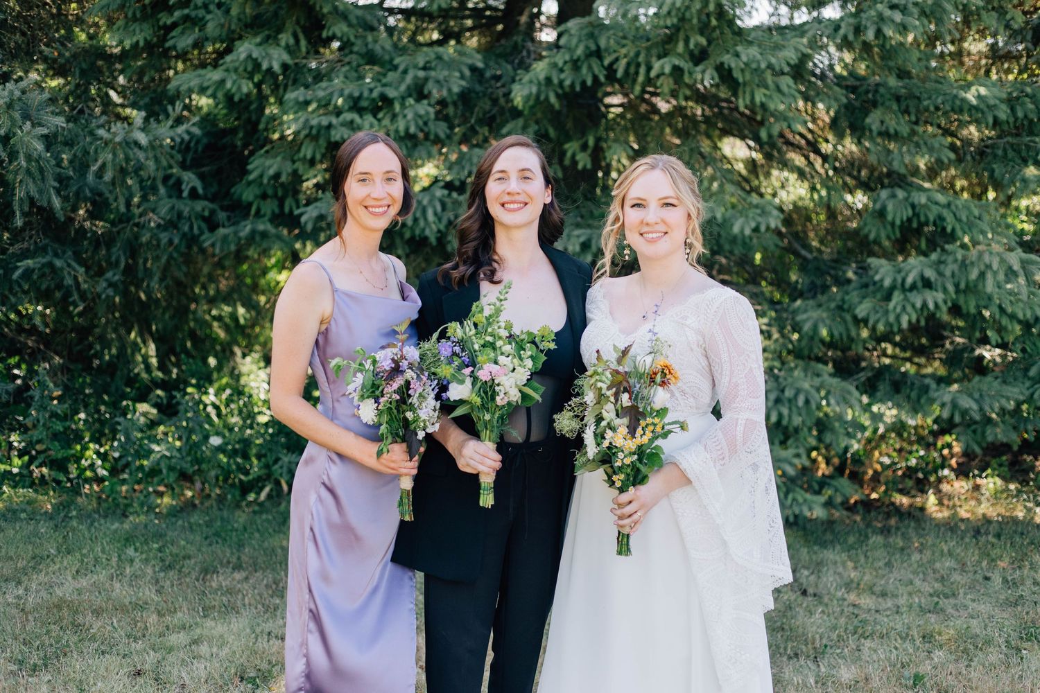 Bride in white gown poses with two attendants holding purple and white bouquets before evergreen trees.