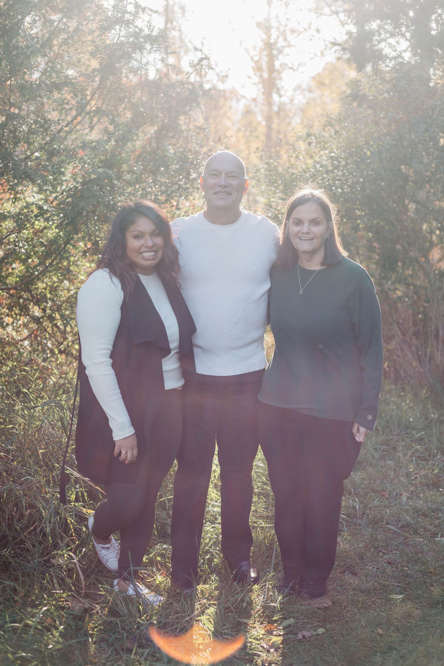 Three adults smiling together outdoors surrounded by golden autumn trees in bright sunlight.