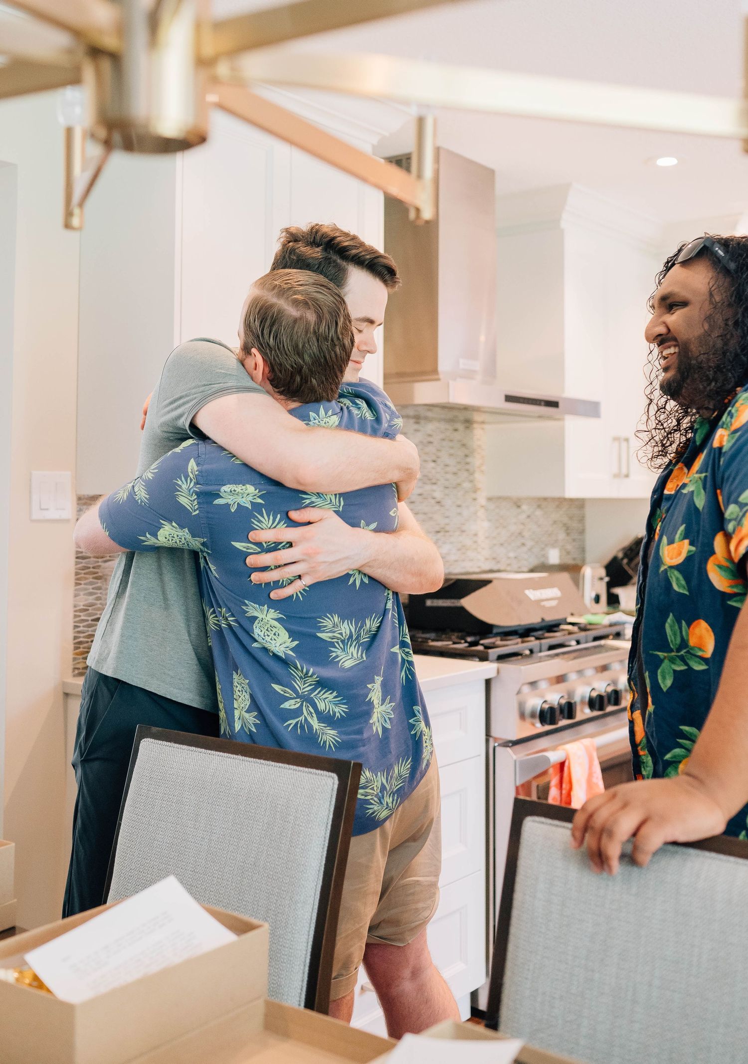 Two men hugging warmly in a modern kitchen while a third man smiles nearby at a wedding gathering.