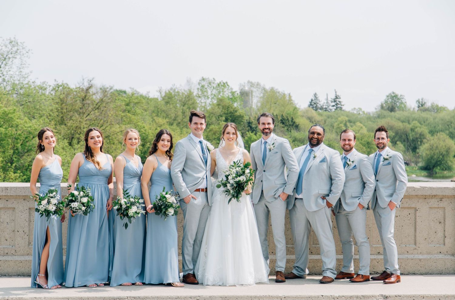Wedding party poses on bridge, bridesmaids in dusty blue gowns and groomsmen in light gray suits with blue ties.