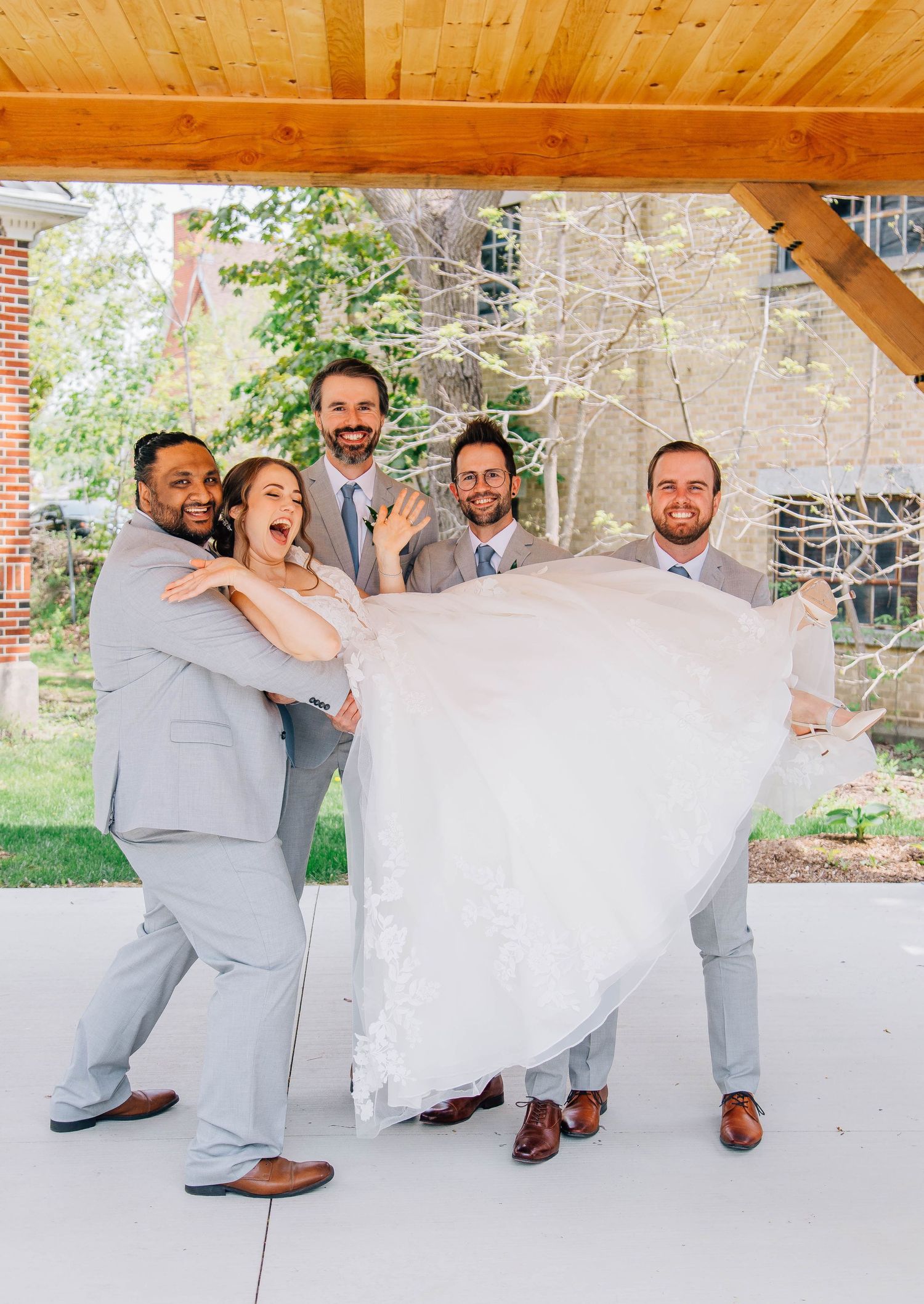 Groomsmen in gray suits playfully lift the bride in a white gown outdoors at a wedding celebration.