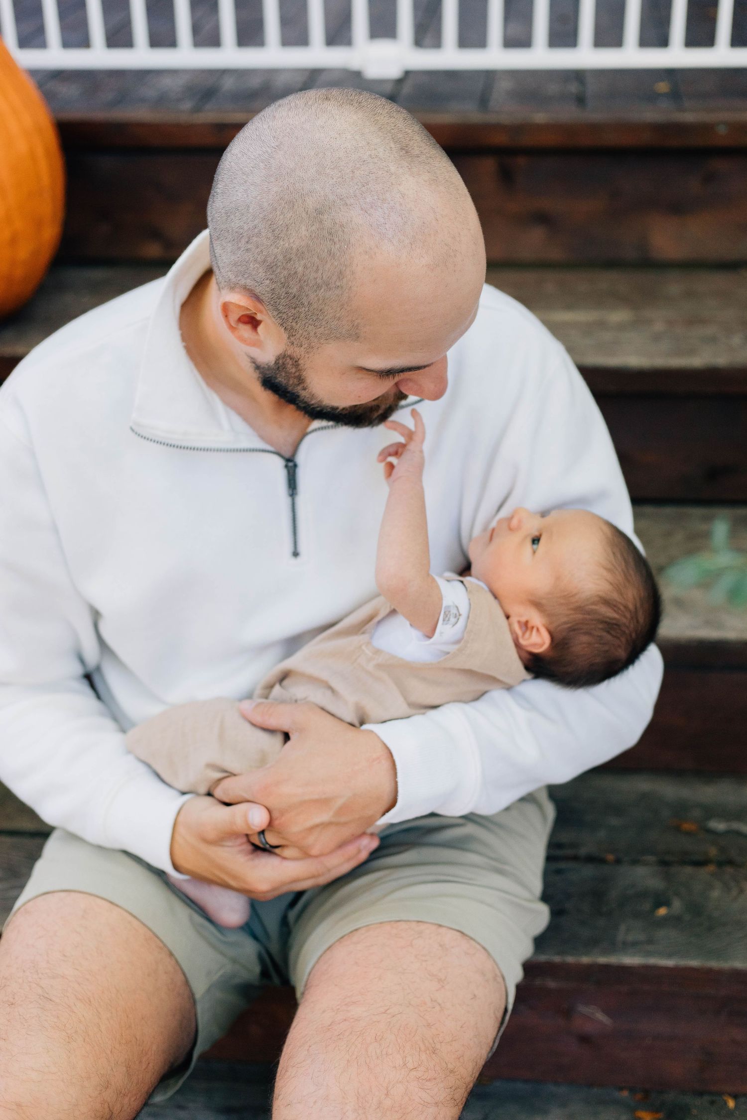 Father sitting on wooden steps holding newborn baby while gazing lovingly at infant outdoors.