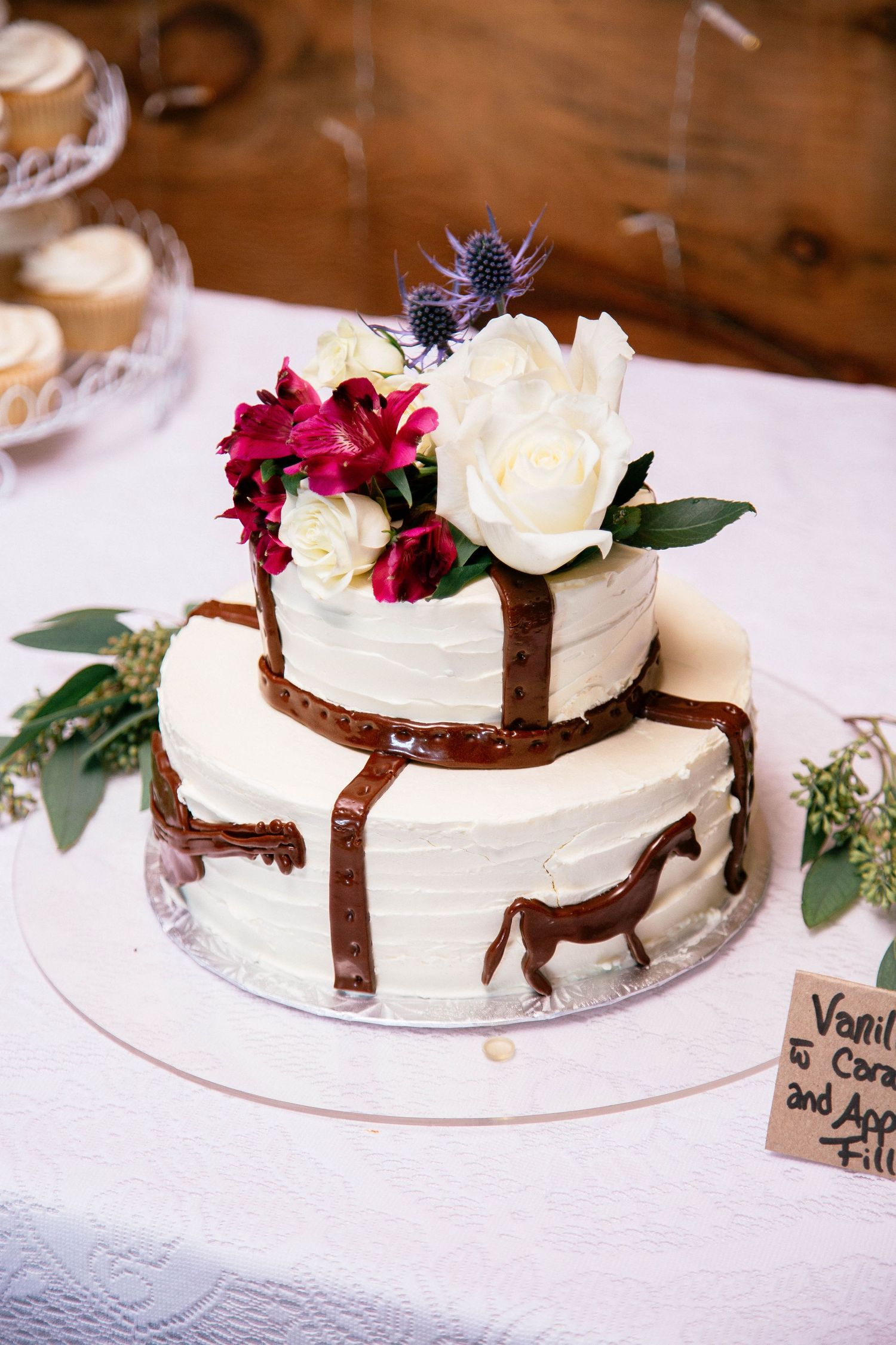 Two-tier white wedding cake with chocolate drip, horse figurine, and fresh white and pink flower topper.