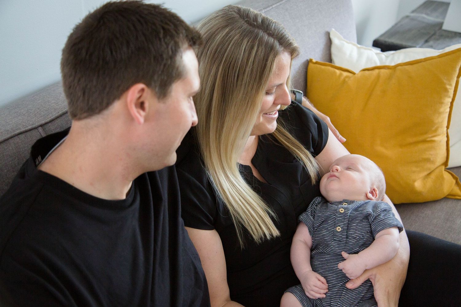 Parents in black shirts smiling at alert newborn baby on gray sofa with yellow pillow.