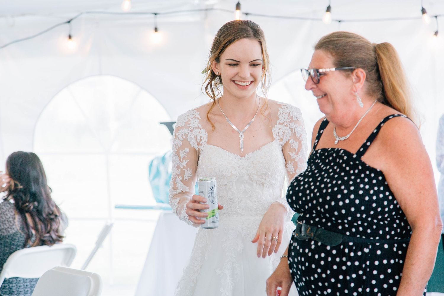A smiling bride in lace gown holds a drink while chatting with a guest in polka dots at her reception.