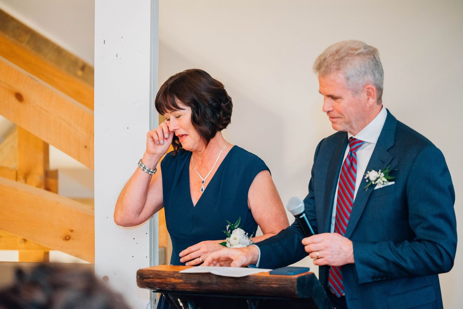 Emotional mother wiping tears while giving wedding speech at podium with father of bride beside her.