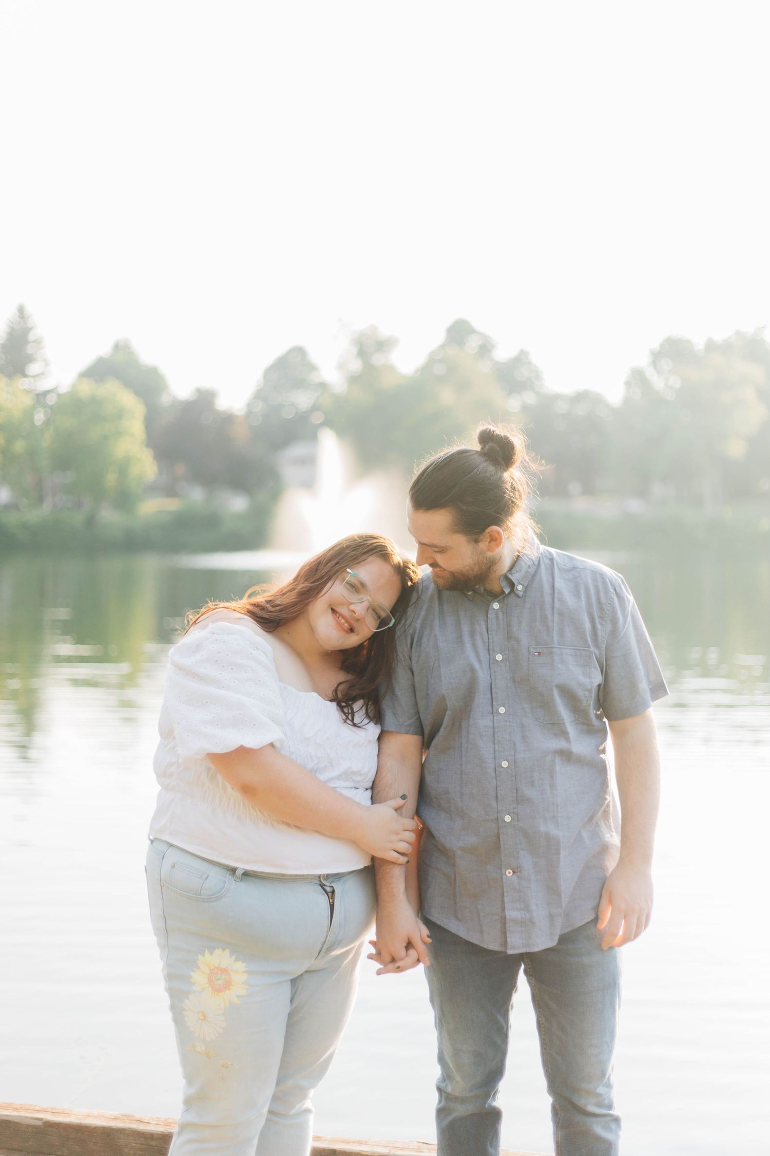 Couple holding hands and smiling by a lake with a fountain, woman leaning on mans shoulder outdoors.