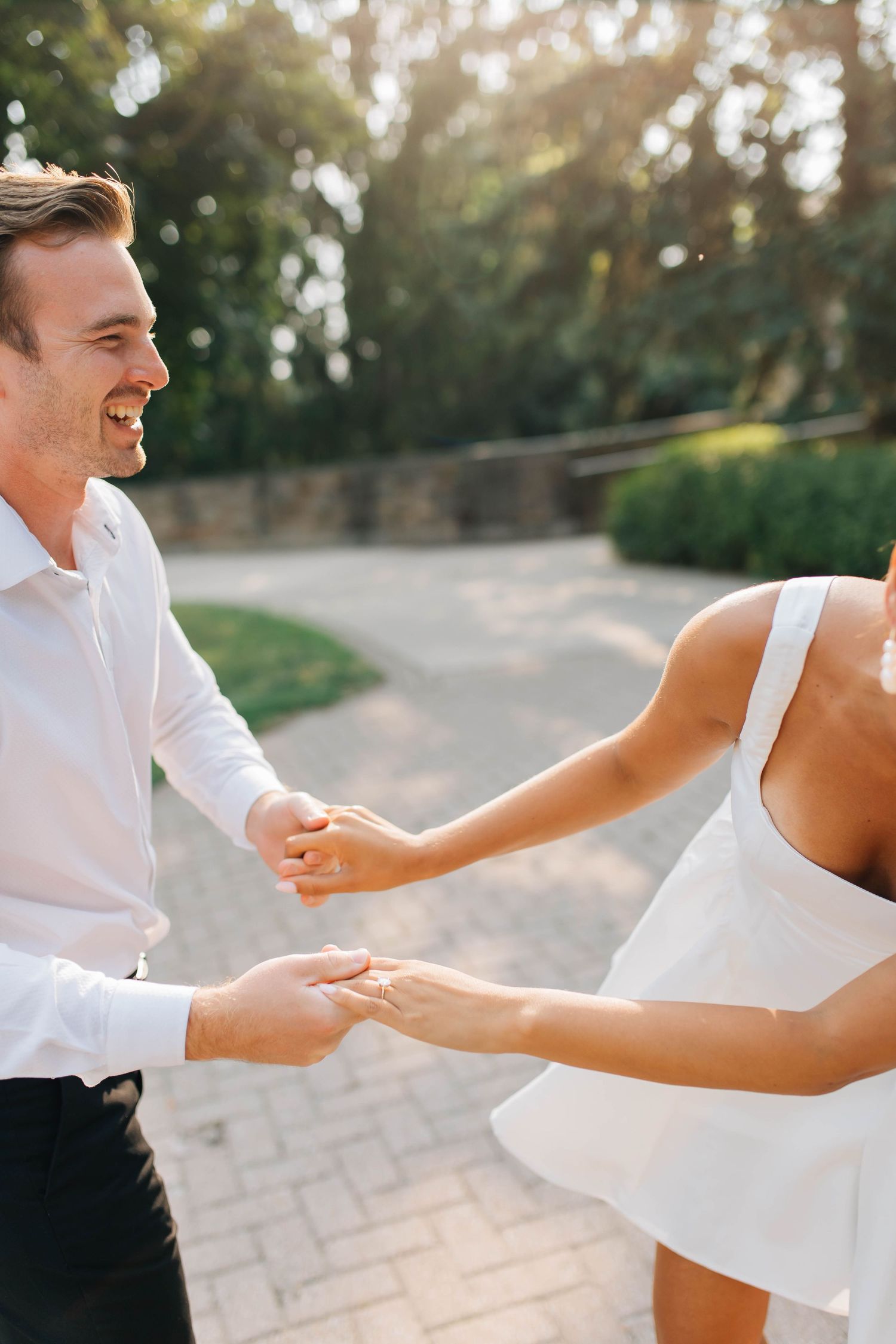 Smiling groom in white shirt holding hands with bride in white mini dress, dancing joyfully outdoors.