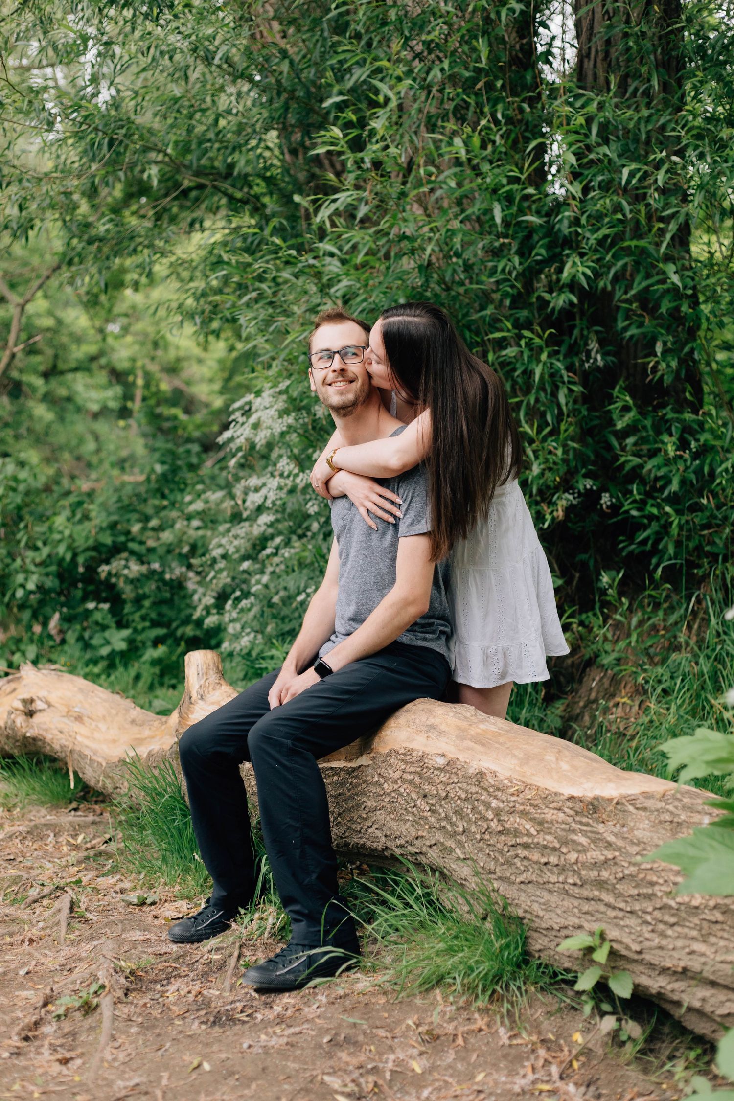 Woman kissing mans cheek as he sits on stone bench surrounded by lush greenery.