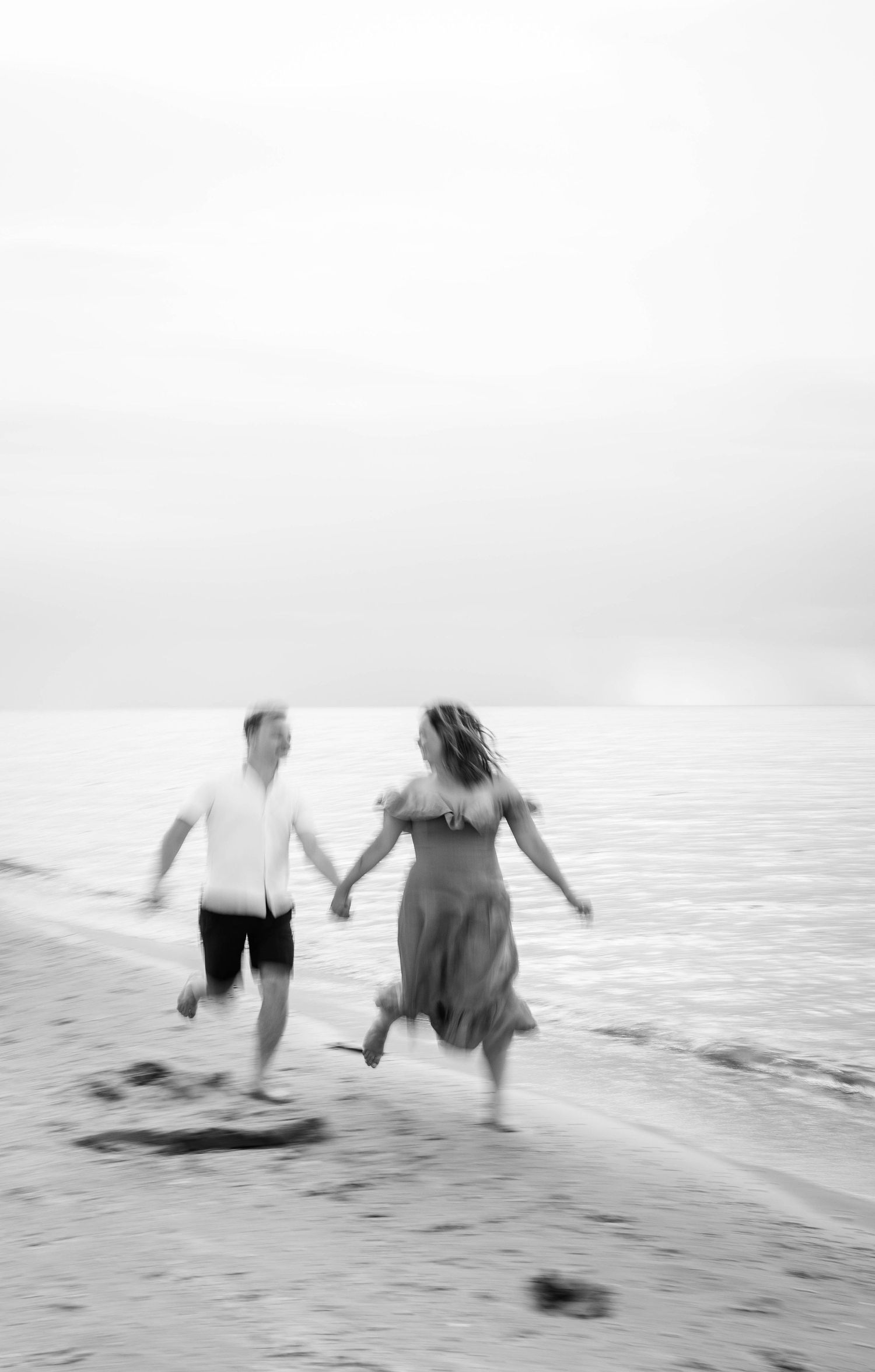 Black and white photo of couple running hand in hand along shoreline, waves crashing at their feet.