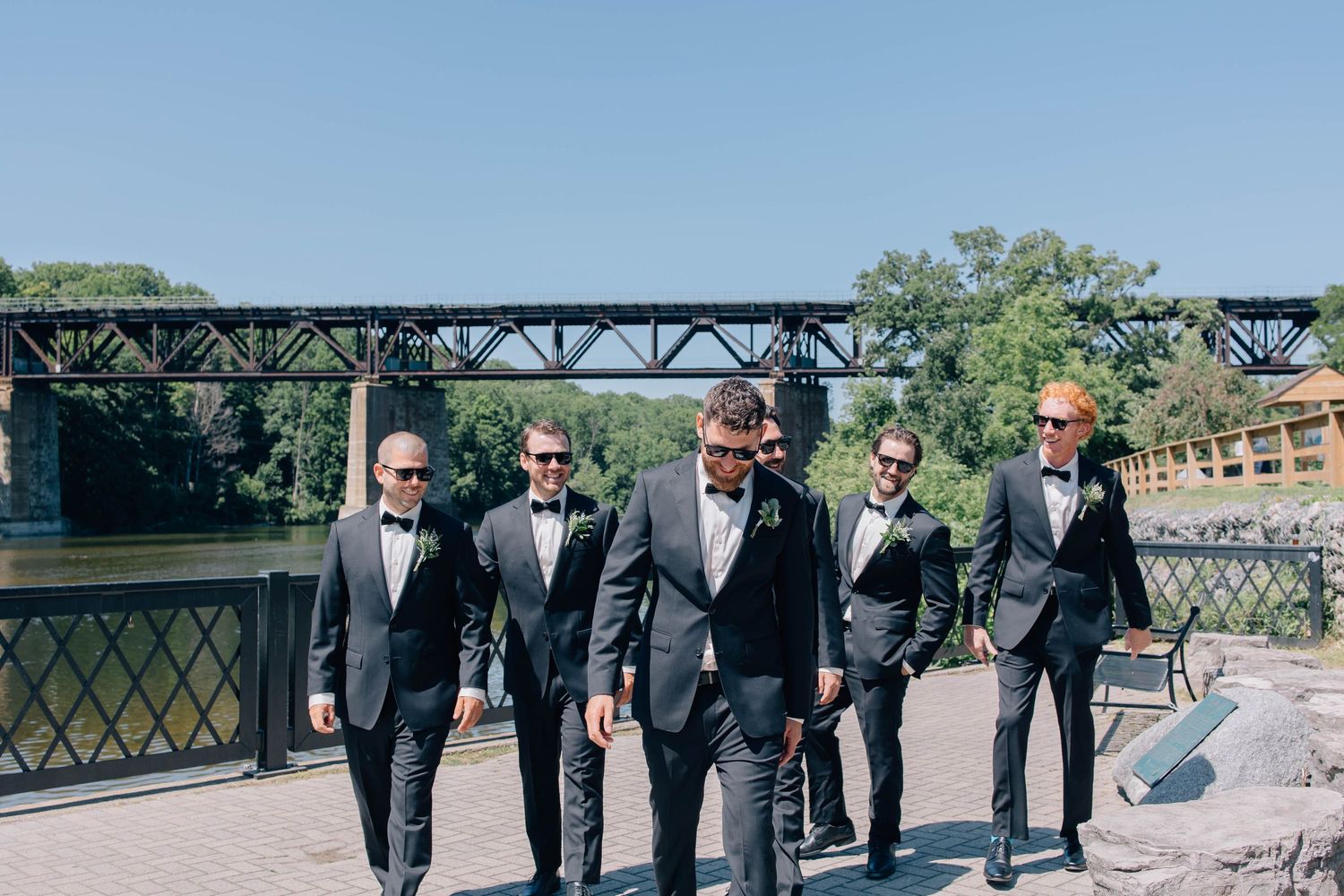 Six groomsmen in black tuxedos walking confidently along a riverside path with a steel bridge backdrop.