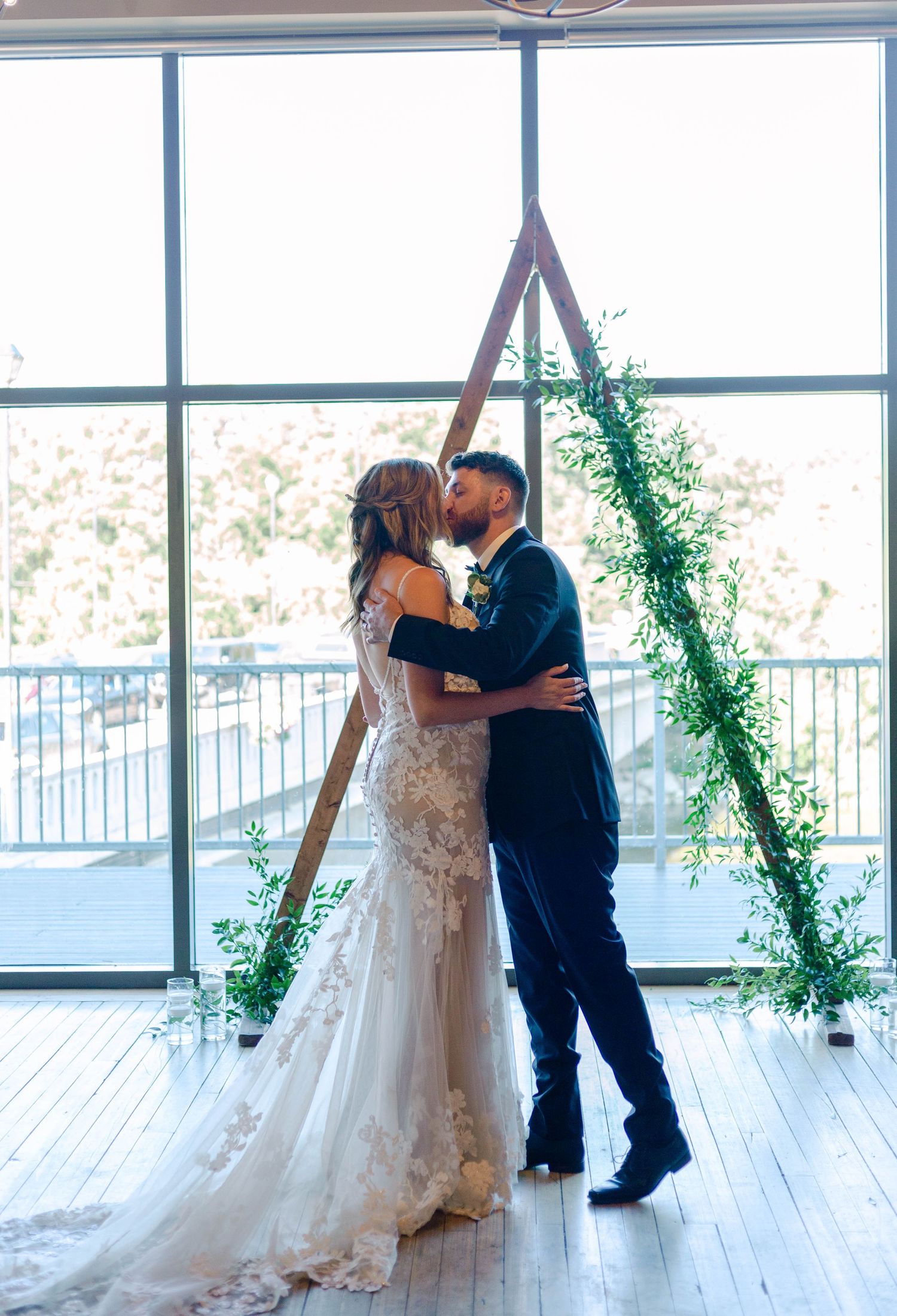 Bride and groom sharing first kiss at triangular wooden arch adorned with greenery in bright windowed venue.