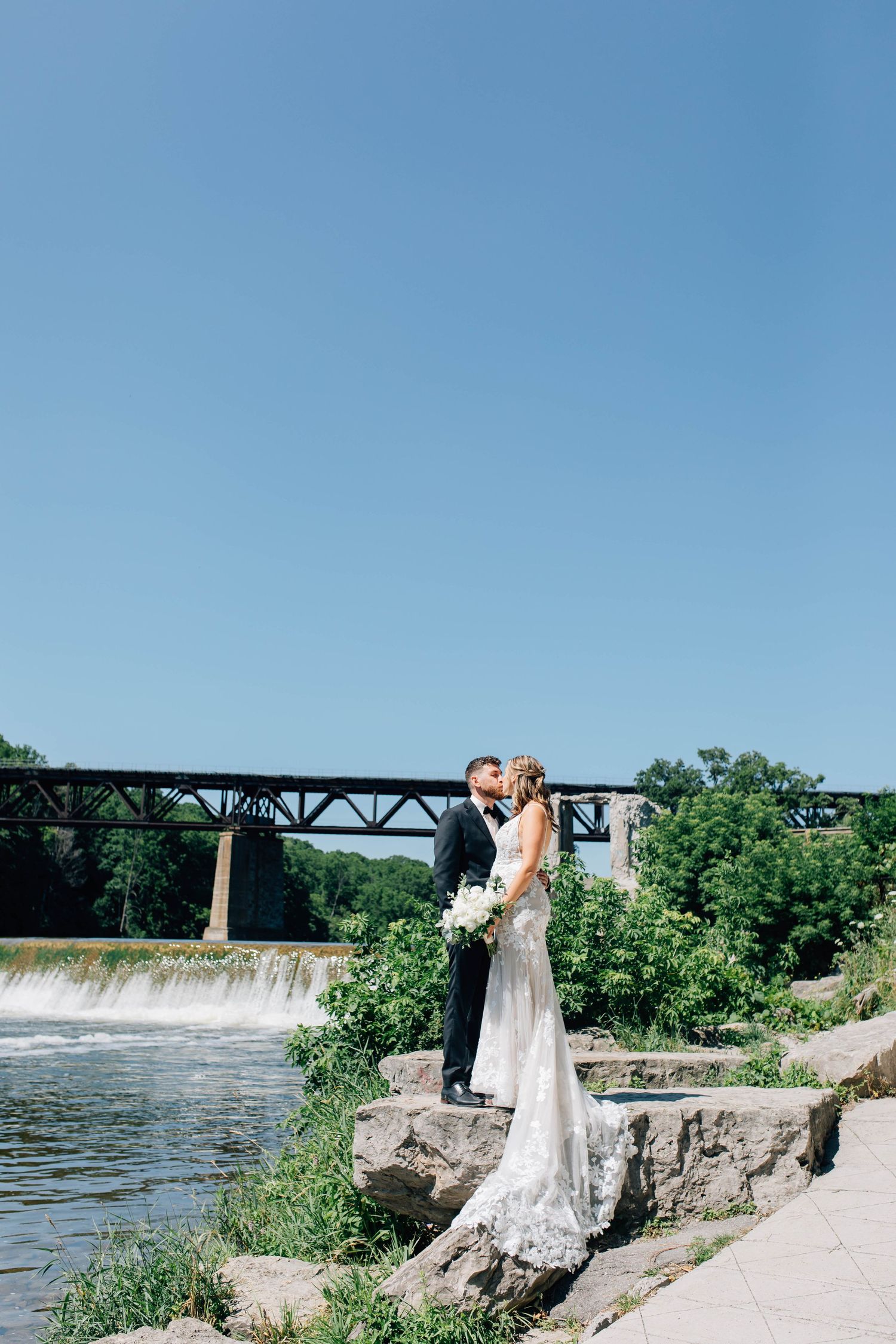Wedding couple sharing a romantic kiss on rocks by a waterfall with an iron bridge in the background in Paris Ontario.