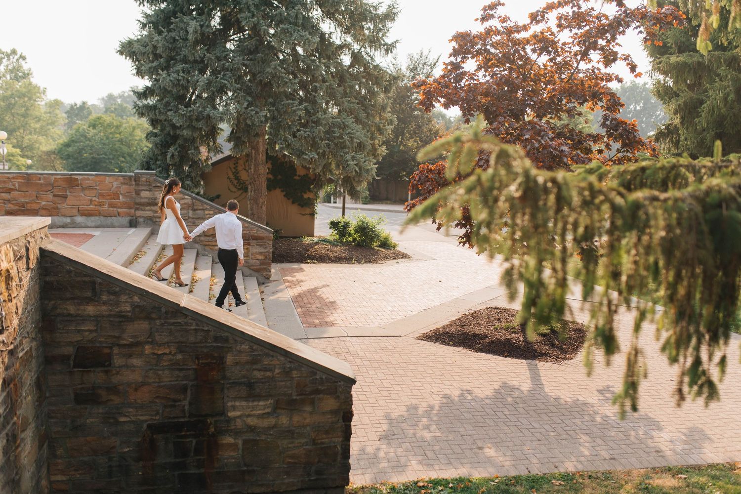 Bride and groom walking hand in hand along a stone wall pathway surrounded by beautiful park scenery.