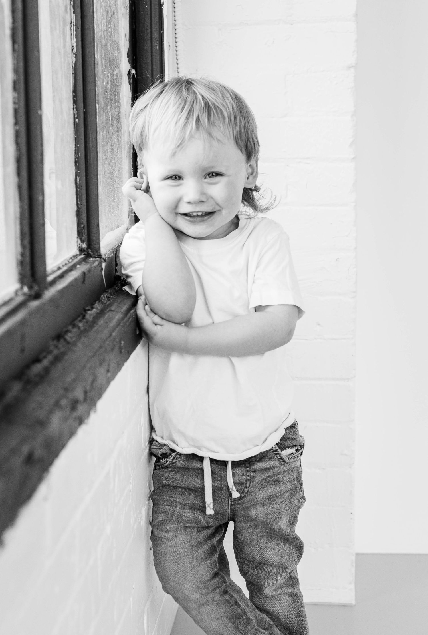 Black and white photo of cheerful toddler boy leaning against window frame smiling at camera.