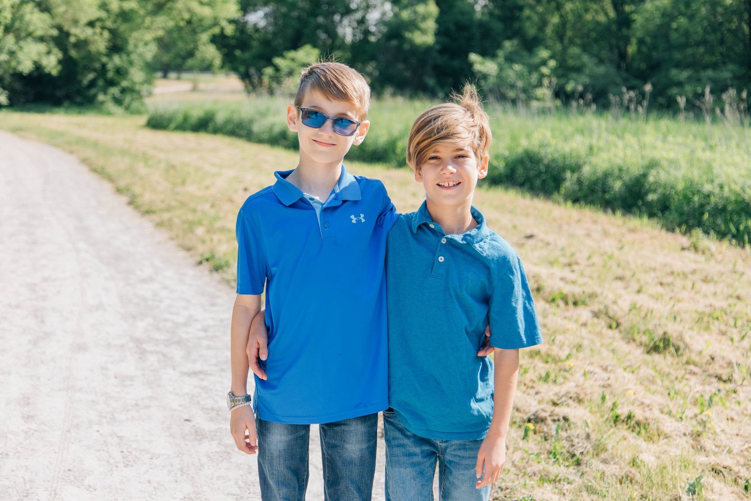 Two smiling boys in blue polo shirts standing together on a sunny gravel path.