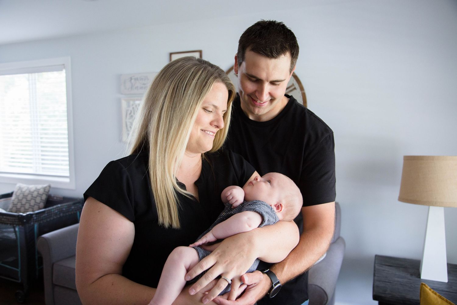 Smiling parents in black shirts cradling their newborn baby together in bright living room.