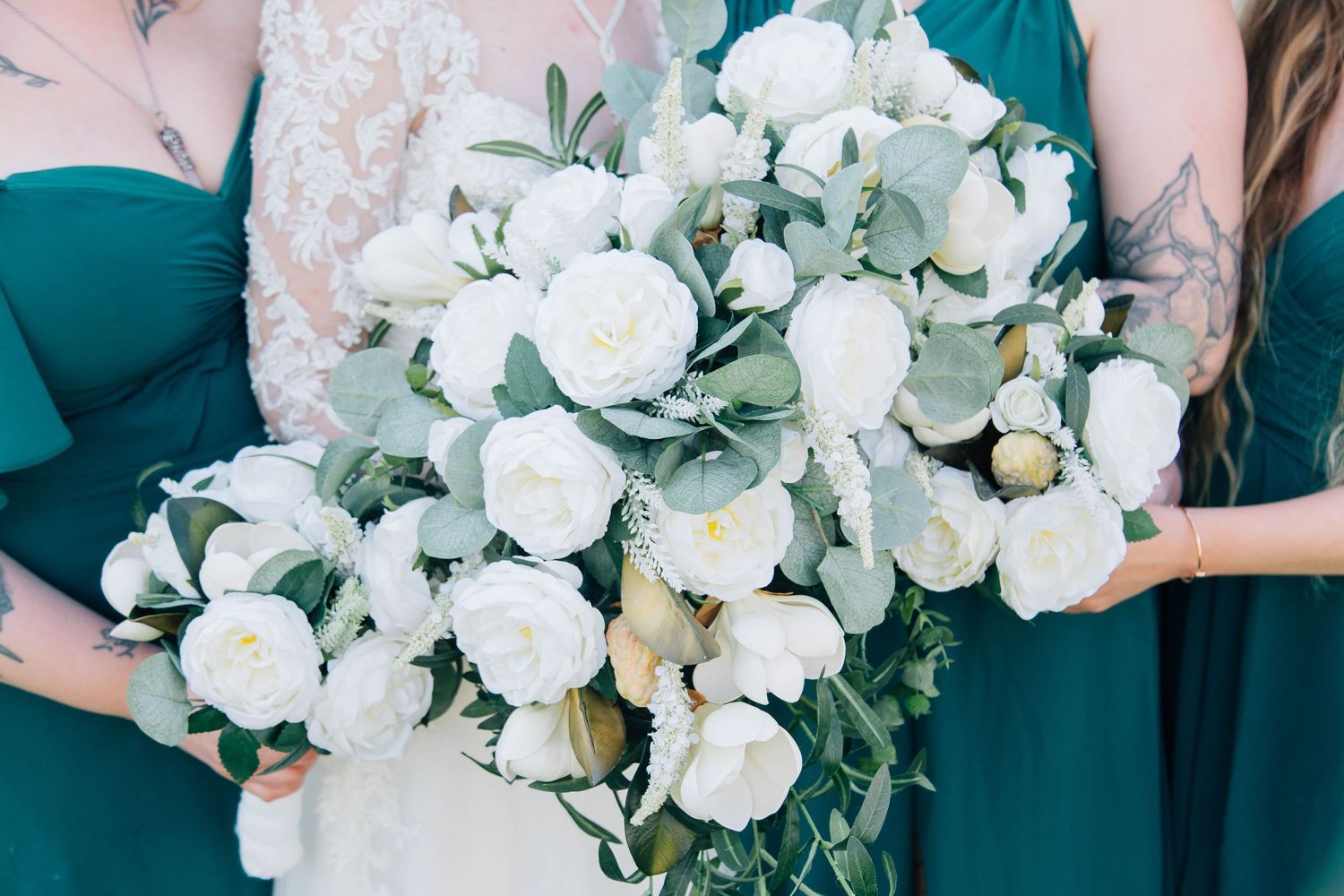 Close-up of a bridal bouquet with white peonies and eucalyptus held by bridesmaids in emerald green dresses.