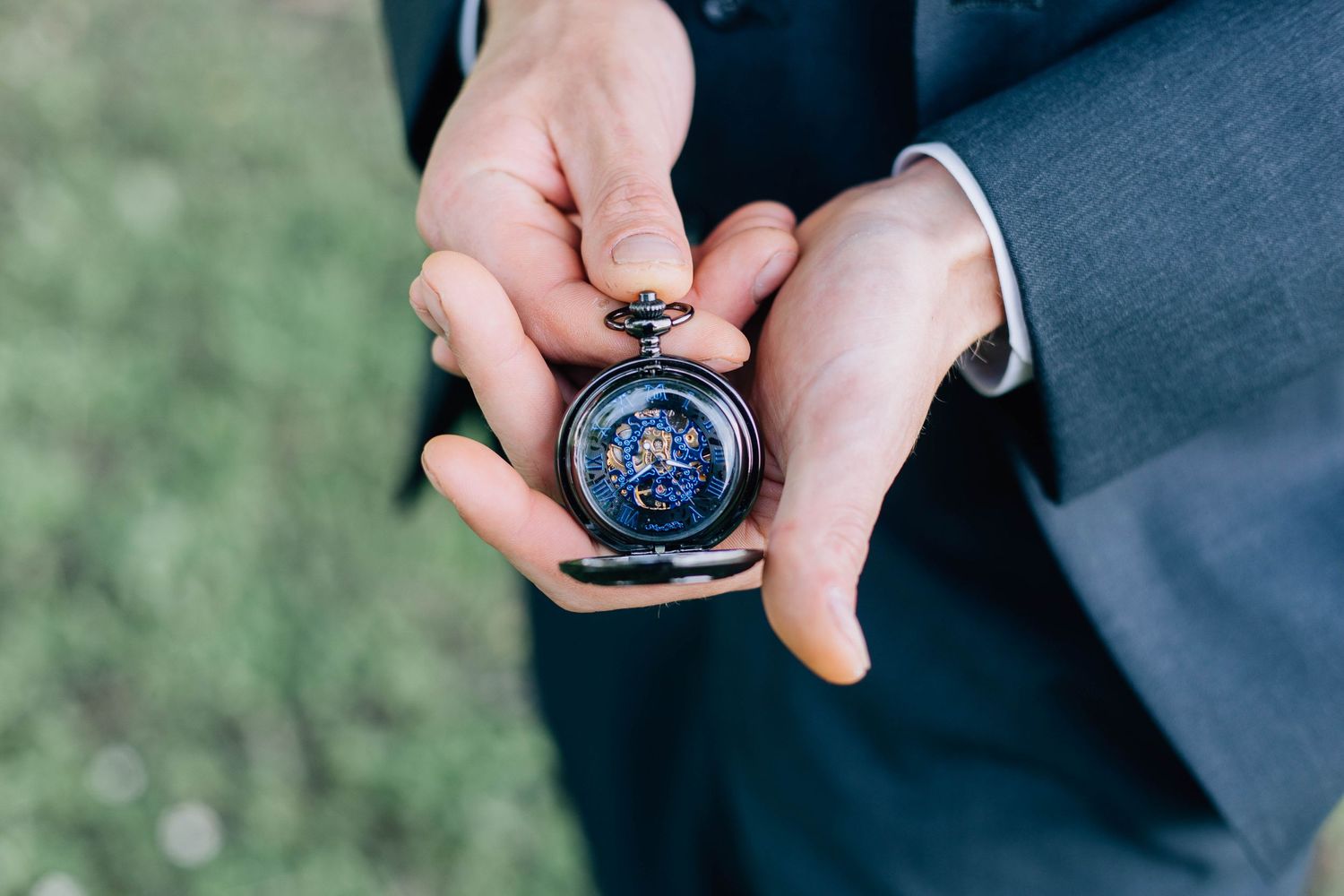 Groom holding an ornate blue celestial pocket watch in his hands outdoors.