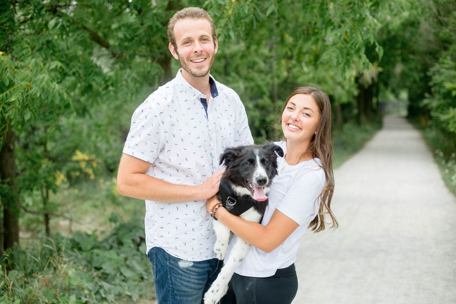 Smiling couple posing with their black and white Border Collie dog on a scenic green nature trail.
