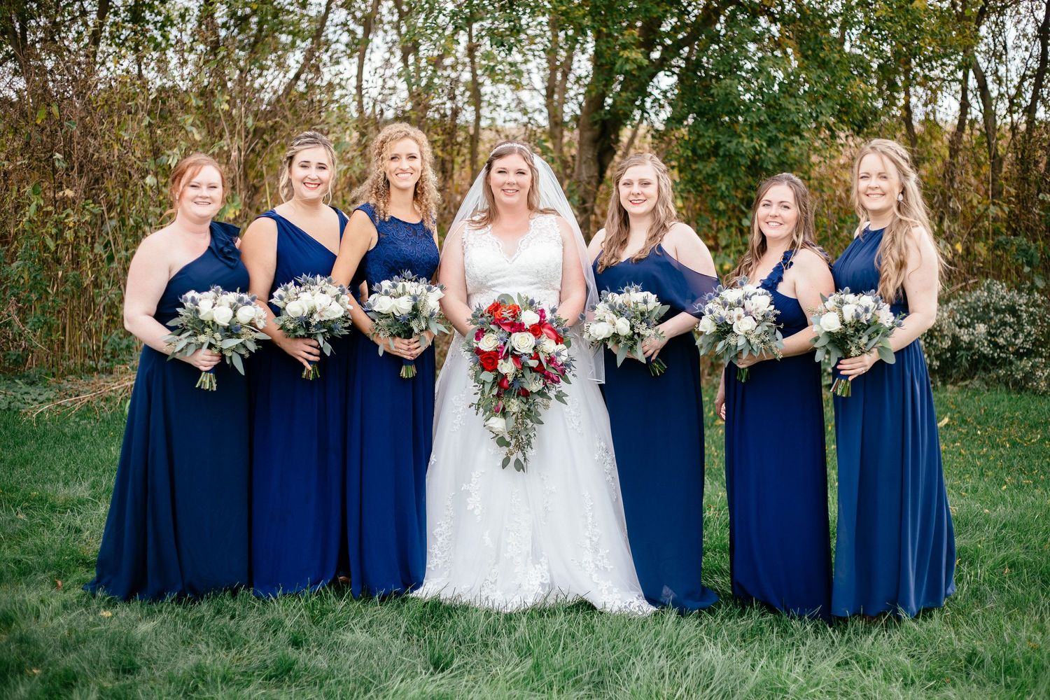 Bride poses with six bridesmaids in navy blue dresses holding white and red bouquets outdoors.