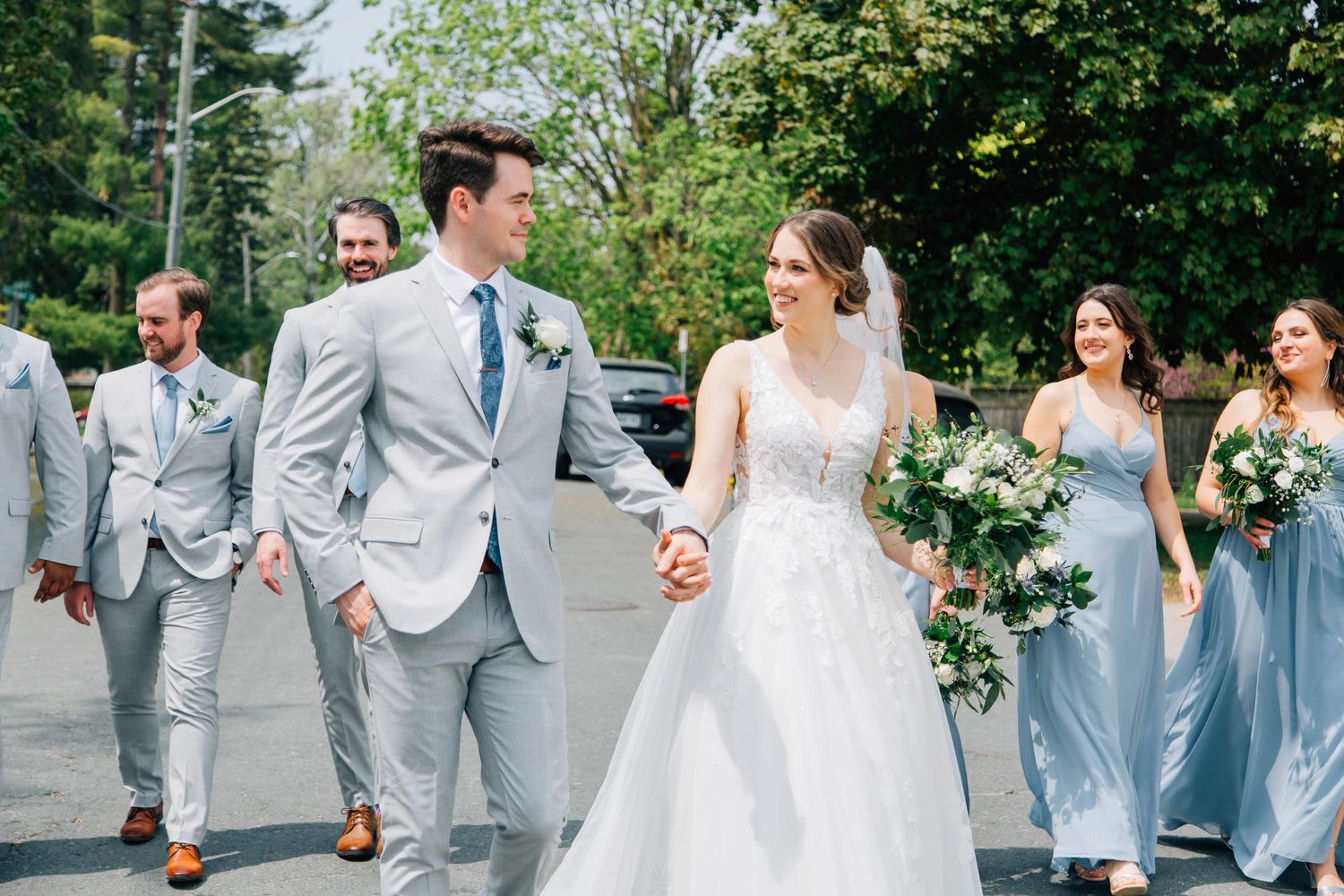 Bride and groom hold hands walking with their wedding party, bridesmaids in dusty blue dresses outdoors.