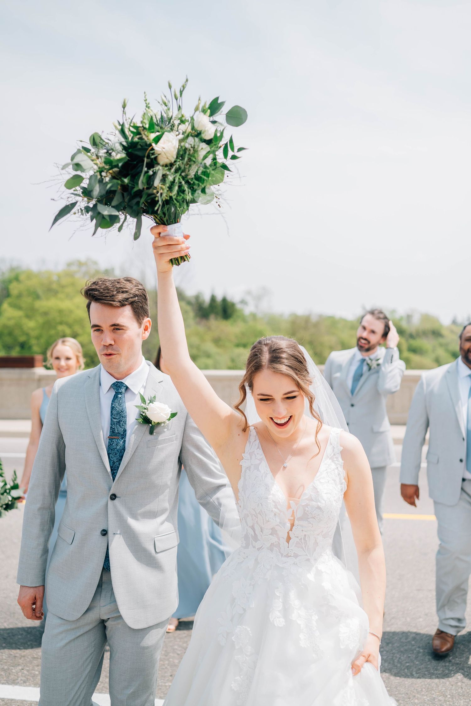 Joyful bride raises white and green bouquet overhead while groom in gray suit walks beside her outdoors.