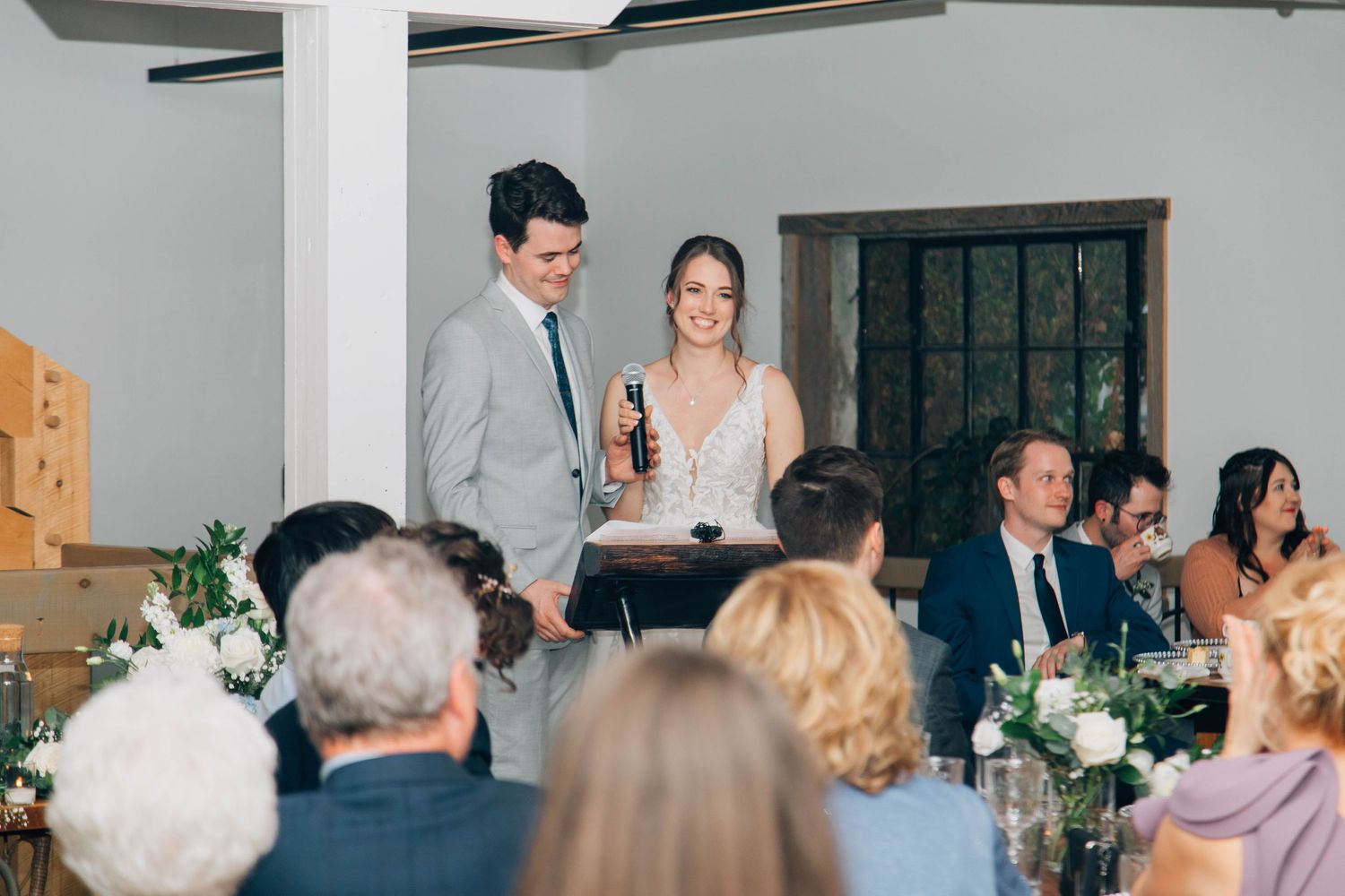 Bride and groom smiling while giving a wedding speech to seated guests in an elegant reception venue.