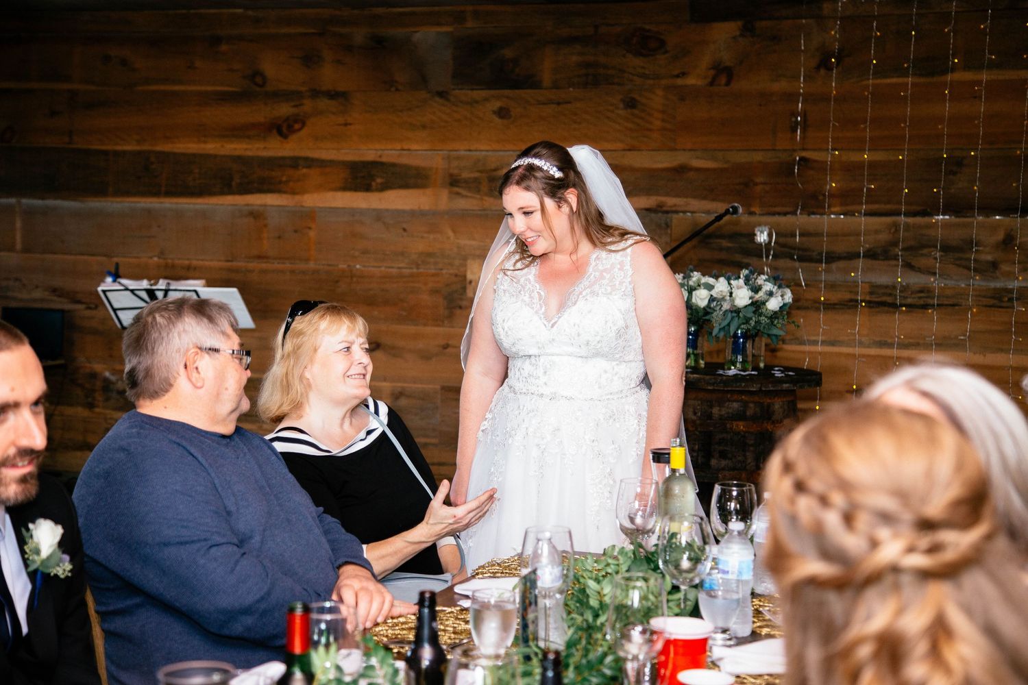 Bride in white gown and veil greets seated guests at decorated wedding reception dinner table.