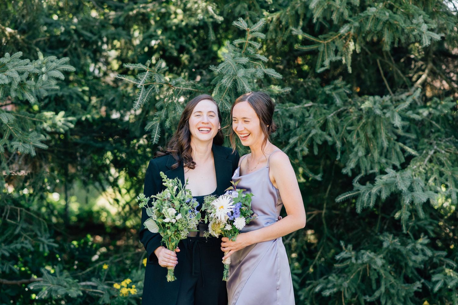 Two smiling women in black and lavender hold floral bouquets together before lush green evergreen trees.