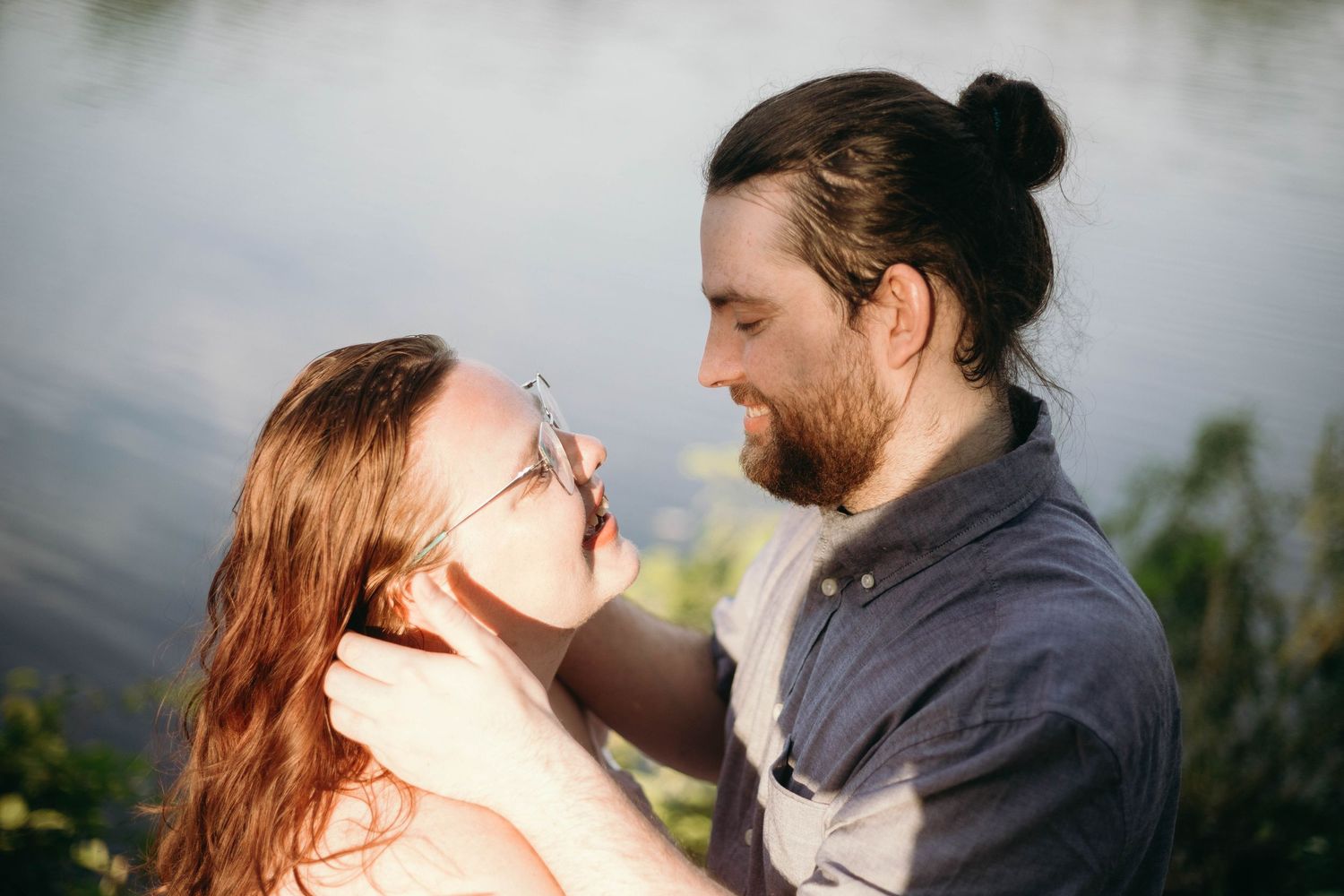 Romantic couple nearly kissing by a lake, man with bun hairstyle and beard smiling at redhead woman.