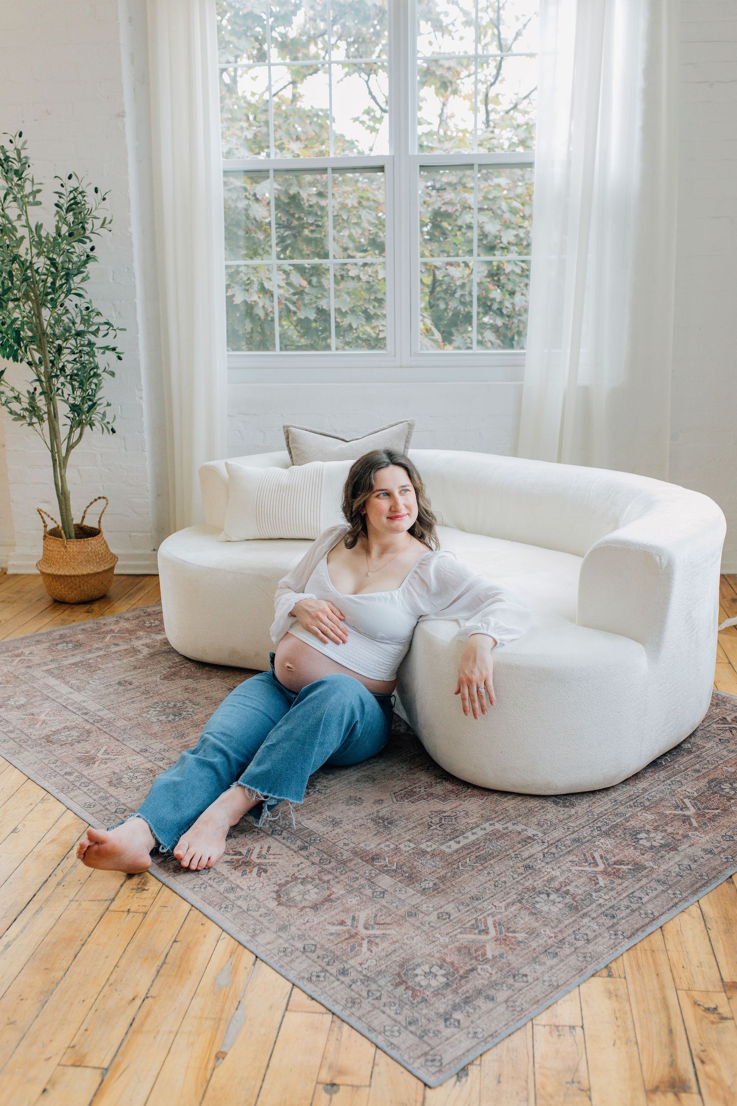 Pregnant woman in white top and jeans sitting on floor leaning against a curved white sofa in a bright living room.