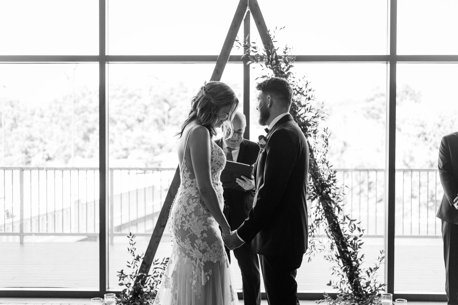 Bride and groom exchanging vows at triangular wooden arch with greenery in black and white wedding photo.