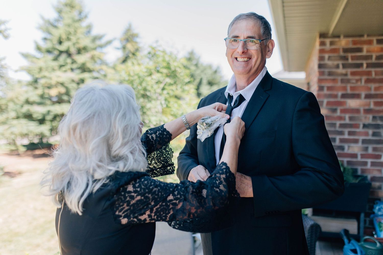 Woman helping smiling man adjust his tie outdoors before wedding ceremony.