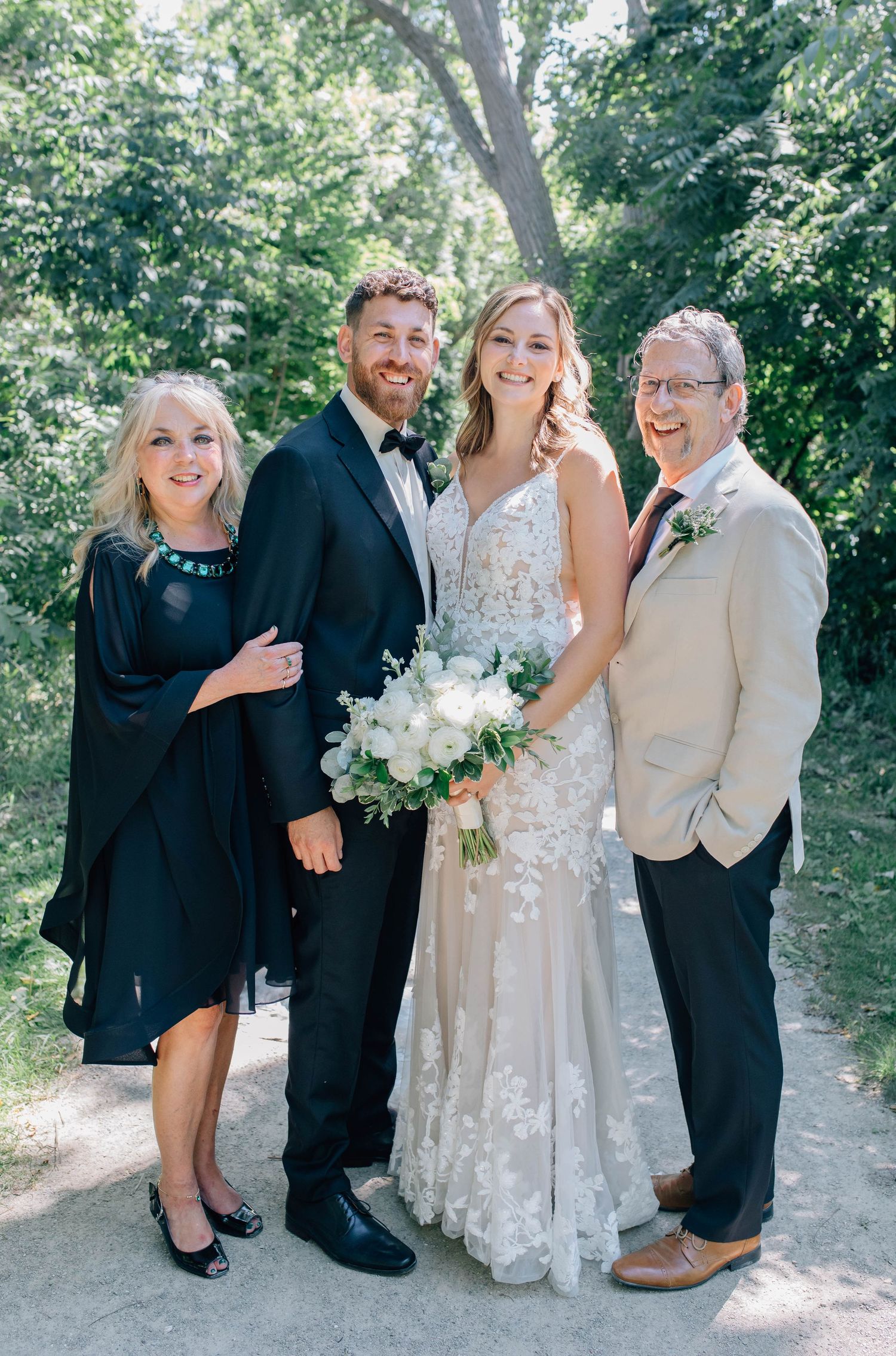 Bride and groom smile with two parents in a sun-dappled outdoor setting surrounded by green foliage.