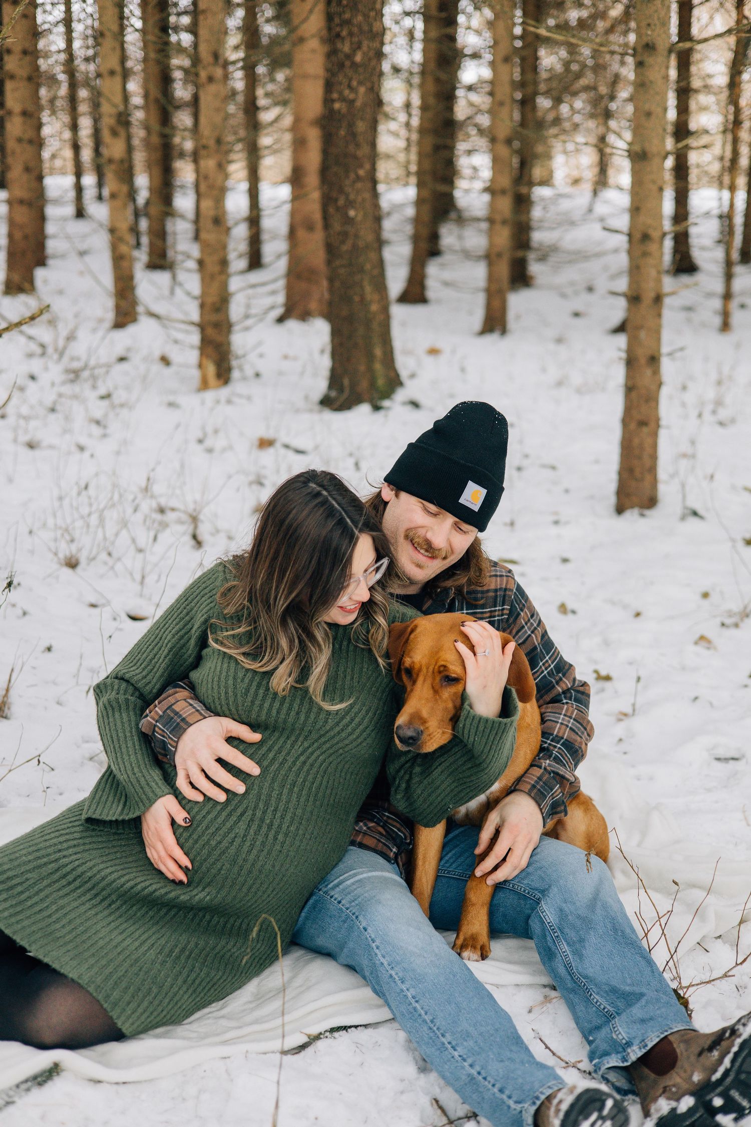 Pregnant couple sitting in snowy winter forest with their brown dog during maternity session.