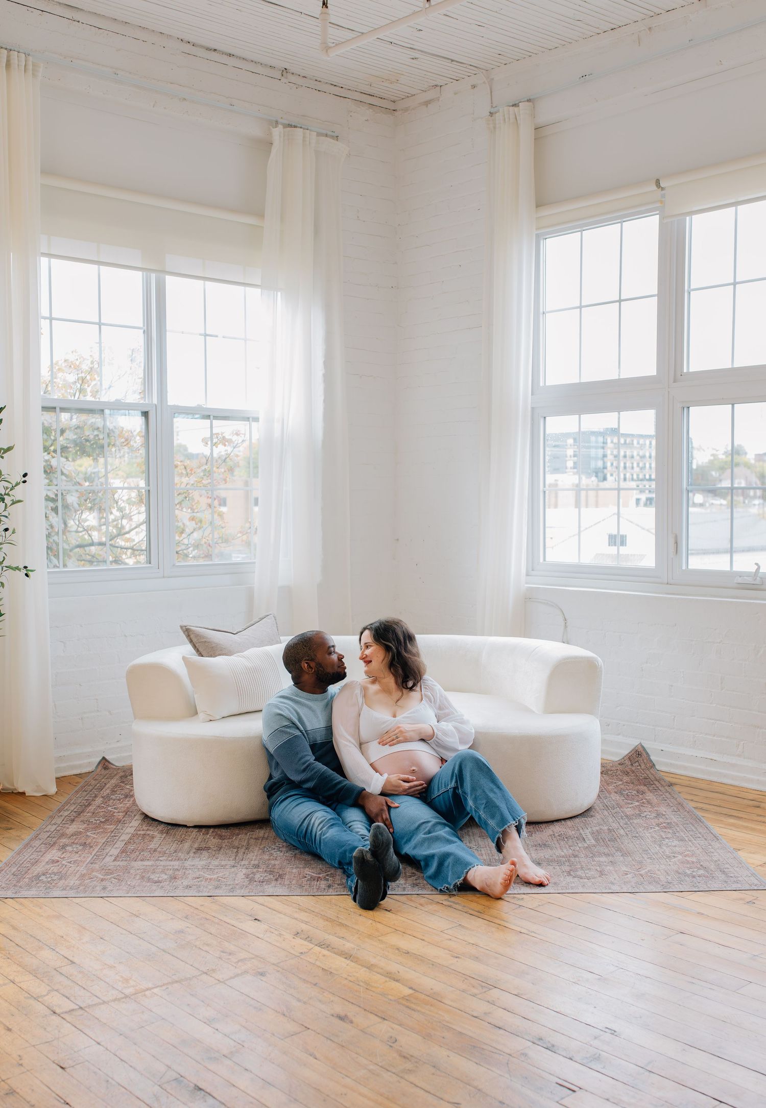 Expecting interracial couple sitting together on floor leaning against white sofa in bright studio.