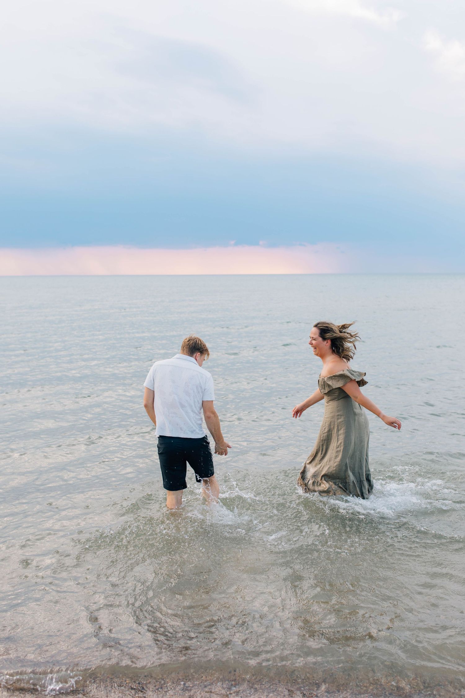 Couple wading through ocean waves at sunset, woman in flowing olive dress splashing in shallow water.