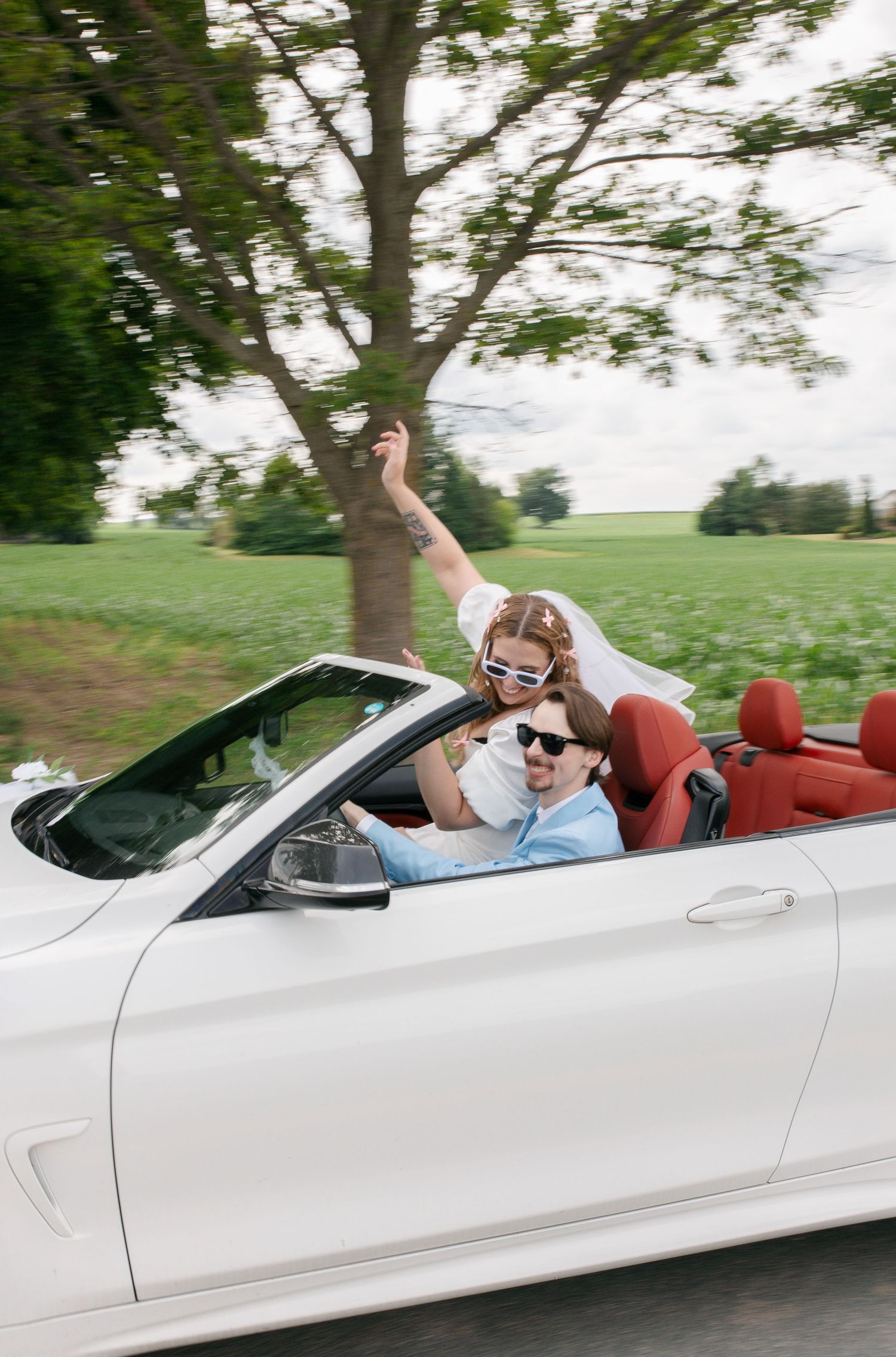A joyful couple in sunglasses riding in a white convertible car with red interior through a scenic road.