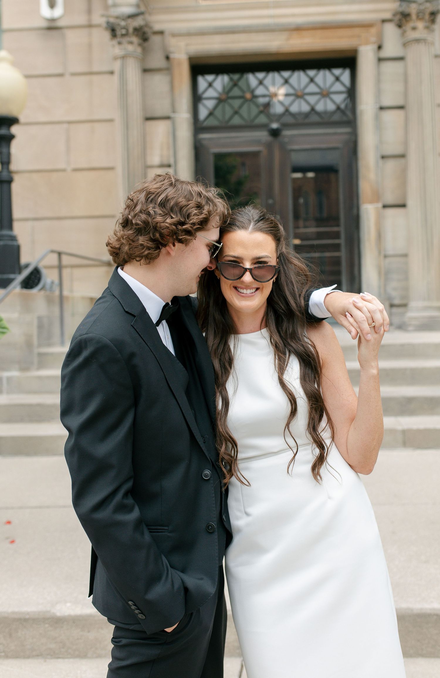 A couple in wedding attire smiles outside a stone building, bride in white dress with sunglasses, groom in black suit.