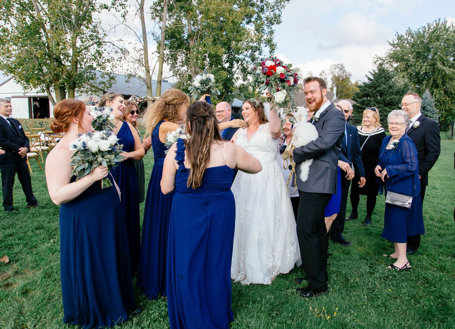 Bridesmaids in navy blue gowns congratulate bride and groom after outdoor wedding ceremony on farm.