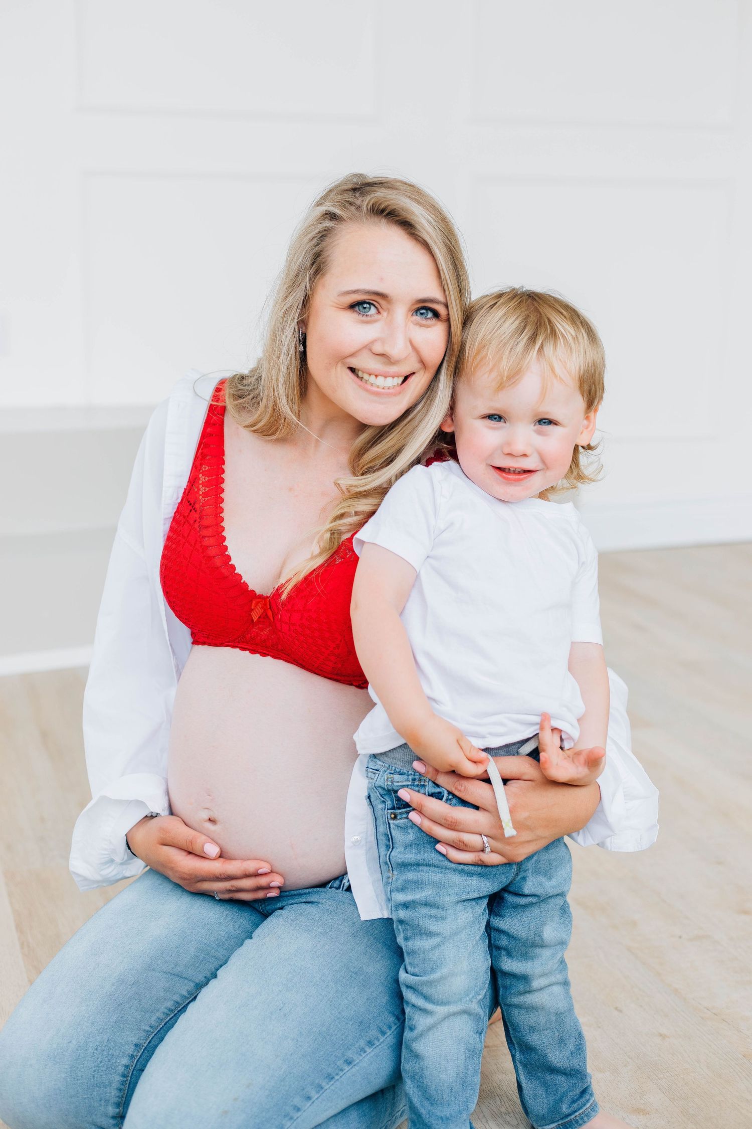 Smiling pregnant blonde mom in red bralette sitting with happy toddler during bright studio maternity session.