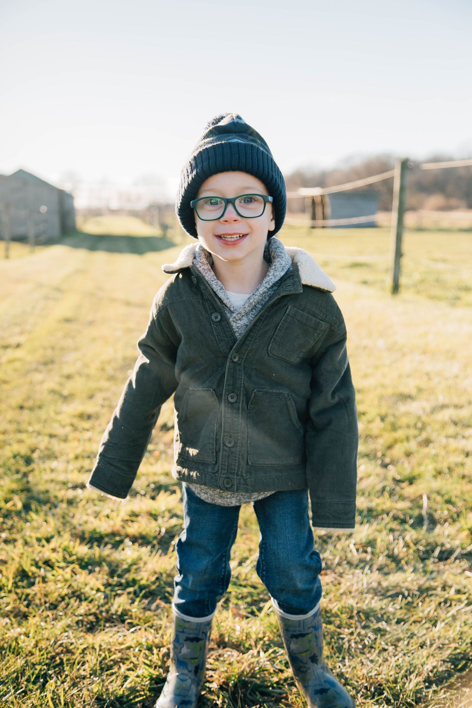 A smiling young boy wearing glasses, a knit hat, and green jacket poses in a sunny farm field.