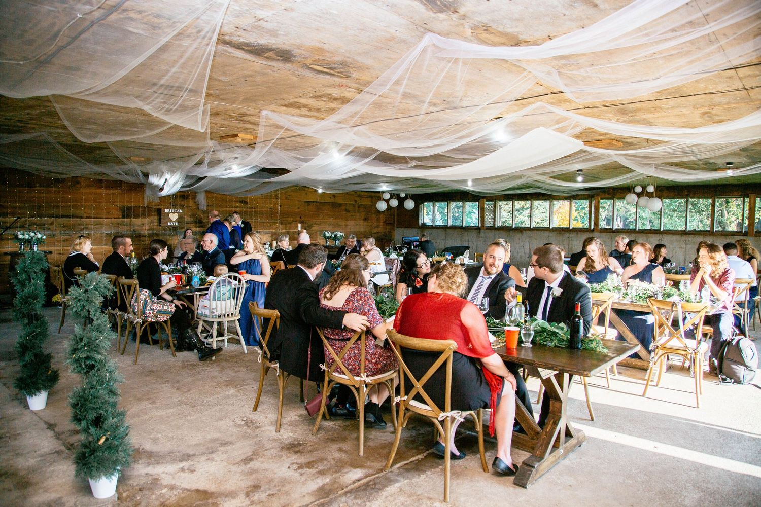 Wedding reception guests seated at rustic wooden tables in a barn venue with draped fabric ceiling decor.