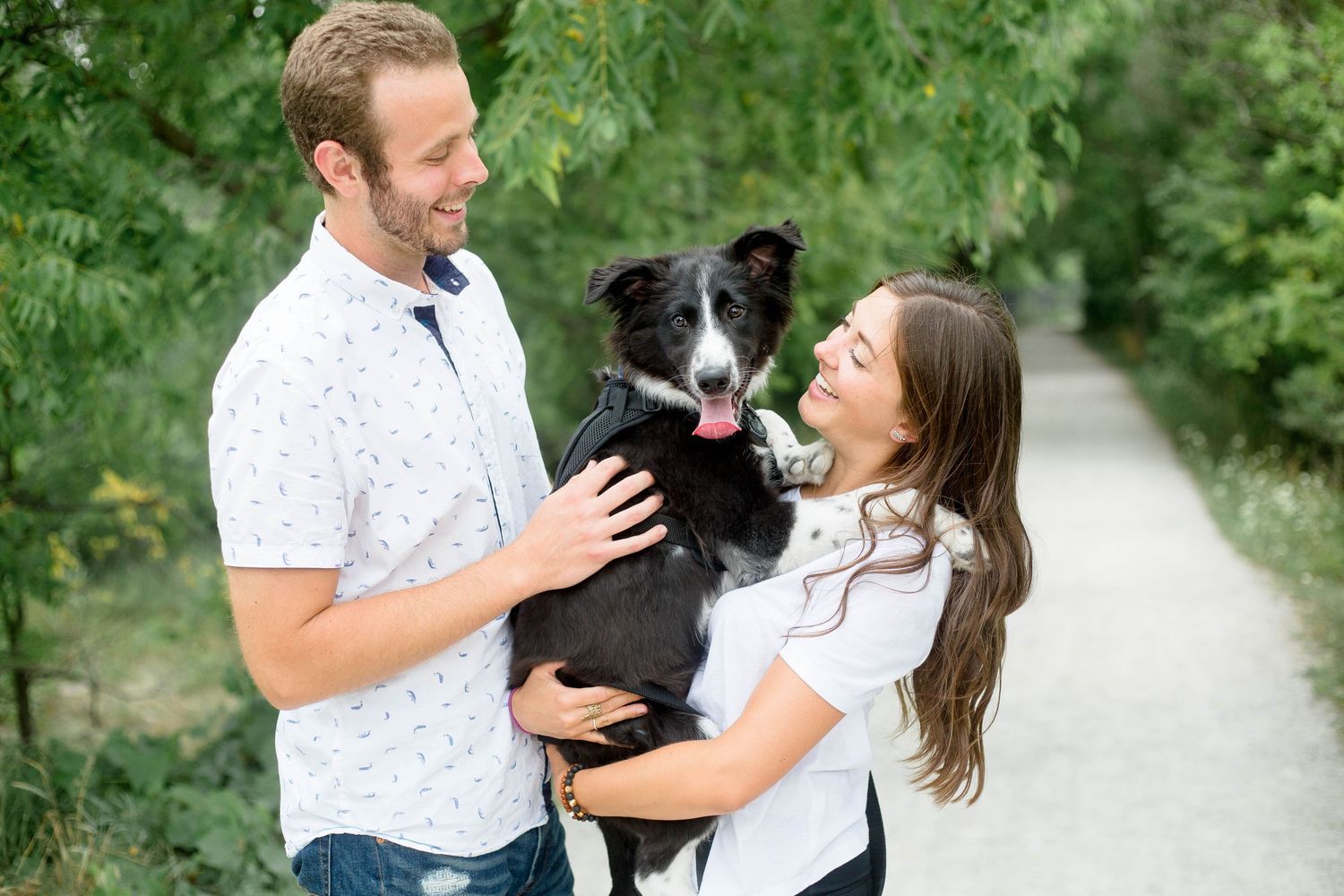Happy couple holding their black and white Border Collie dog together on a lush green tree-lined pathway.
