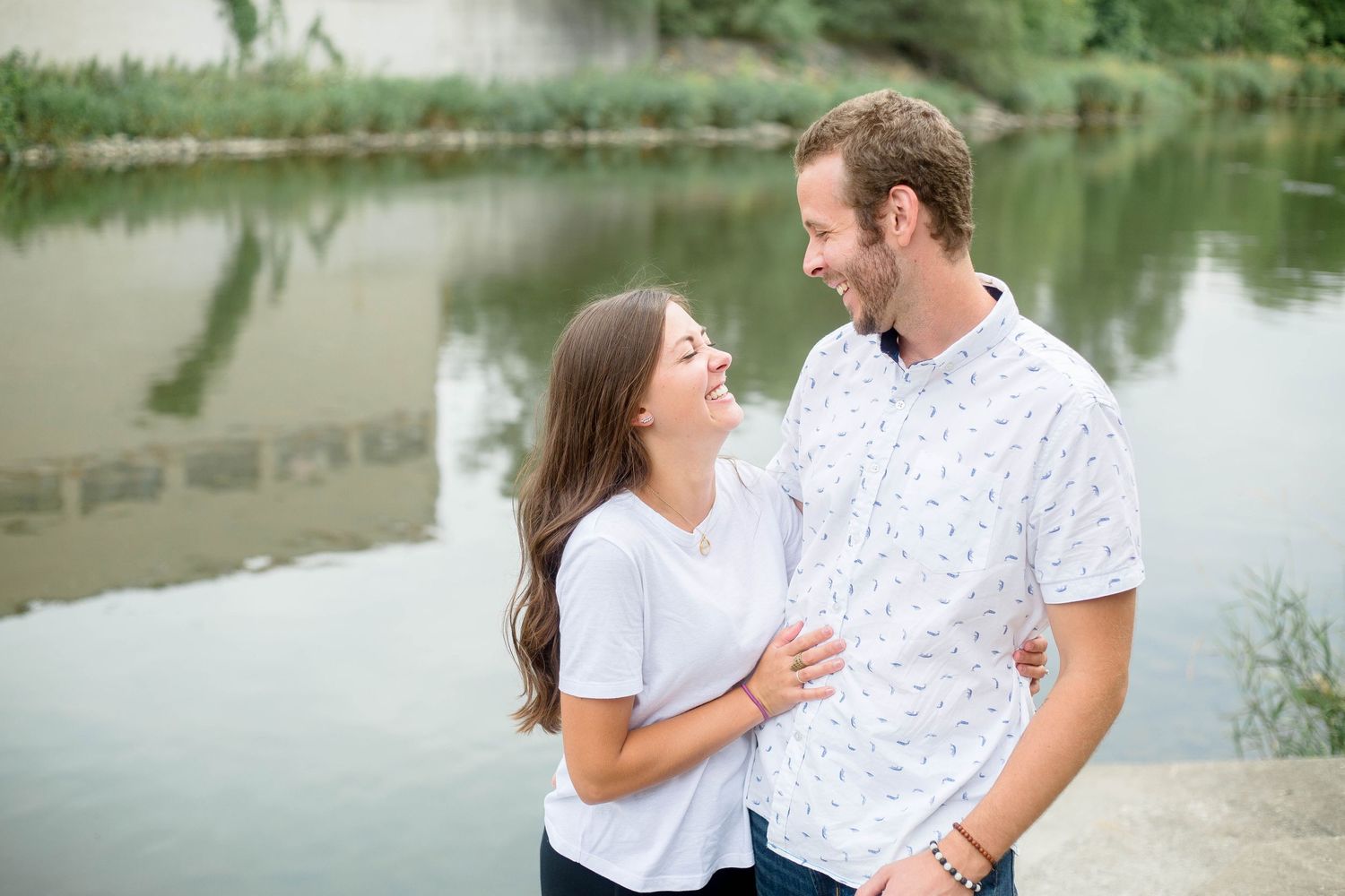A happy couple smiles at each other near a serene pond during an outdoor engagement photo session.